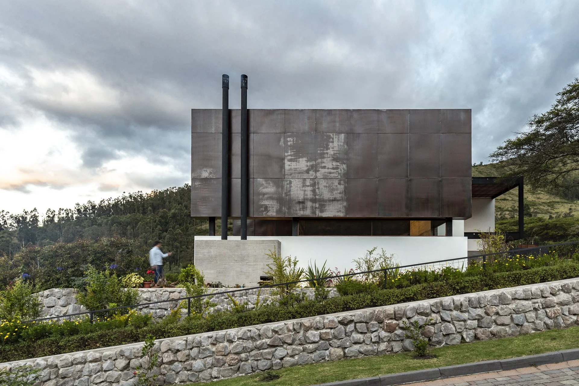 fachada moderna de un edificio con paredes de metal y dos tuberías negras en un entorno natural con árboles y plantas.