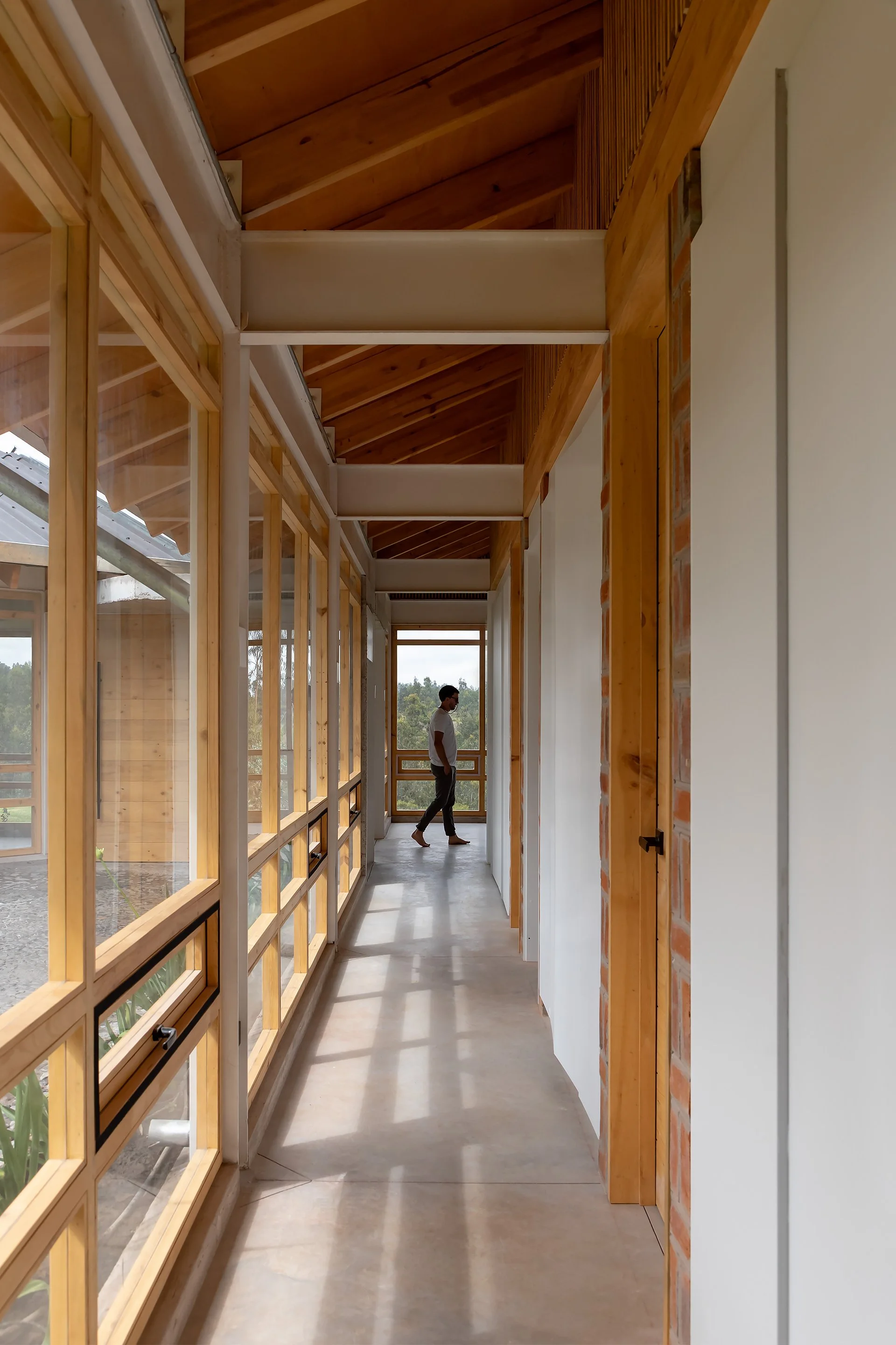 Un pasillo interior con paredes de madera y ventanas grandes enmarcadas en madera. Un hombre de espaldas observa hacia el interior. La estructura del techo también es de madera, con vigas expuestas.