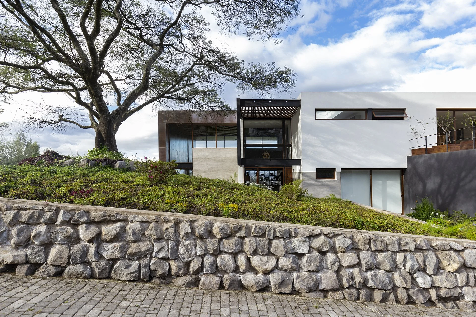 Casa moderna de pisos múltiples, con paredes blancas y ventanas amplias, rodeada de vegetación y un árbol grande en primer plano, bajo un cielo parcialmente nublado.