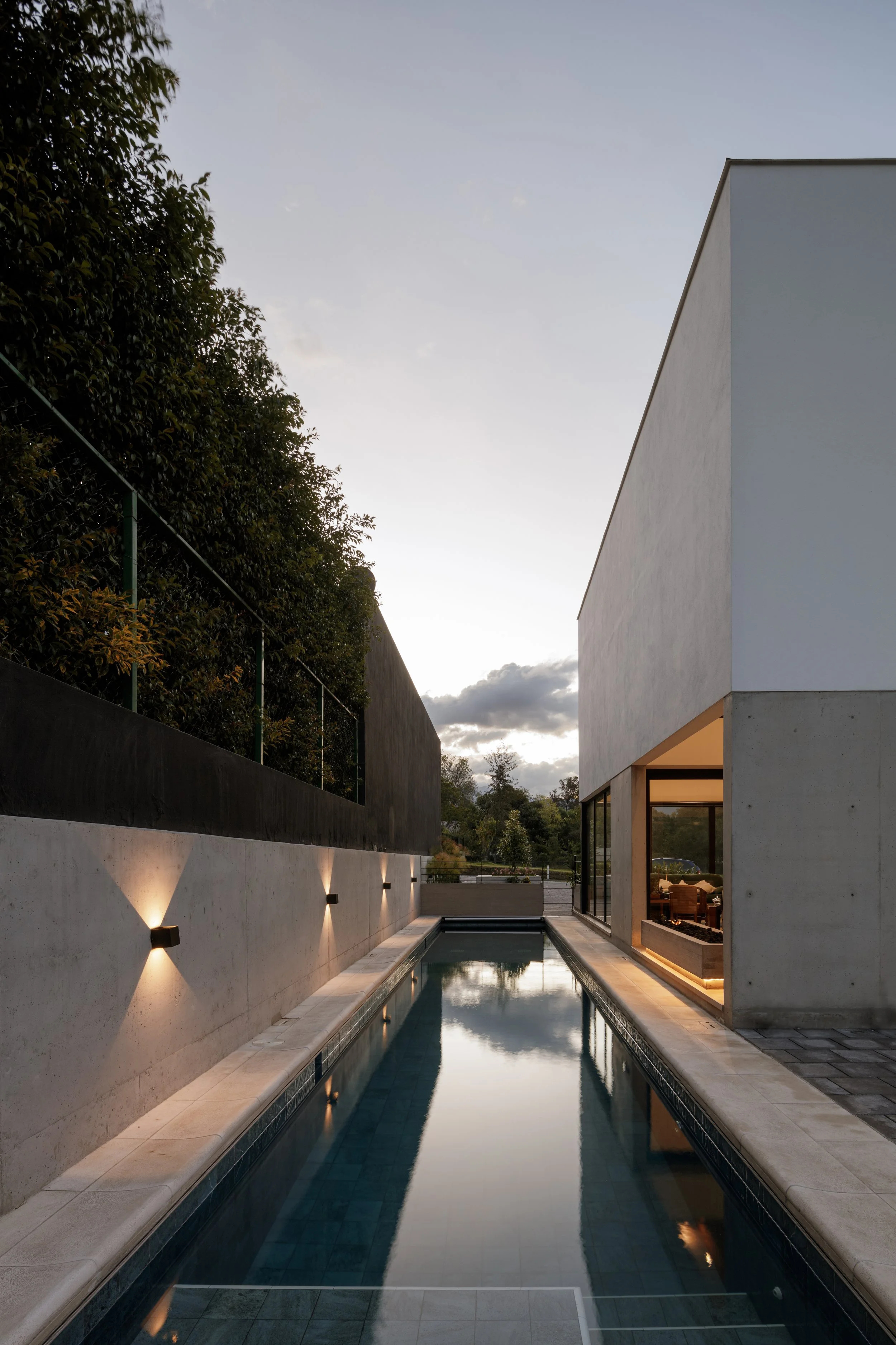 Vista de una piscina estrecha y larga en un área moderna, rodeada de paredes de concreto, con una casa con paredes blancas y ventanas grandes en el fondo, y un cielo nublado al atardecer.