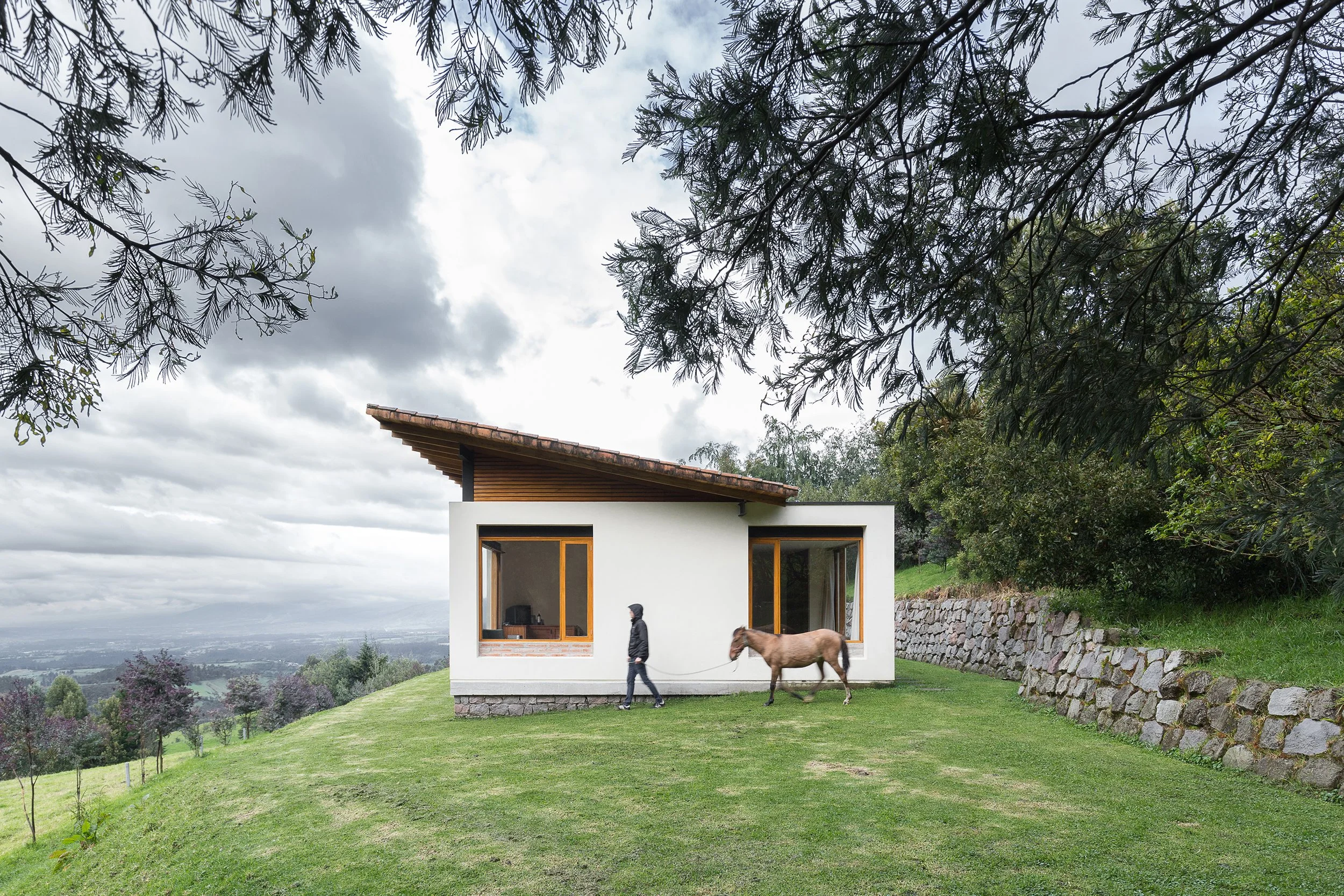 Casa moderna blanca en un entorno rural con árboles grandes y colina con pasto, persona paseando con caballo en primer plano.