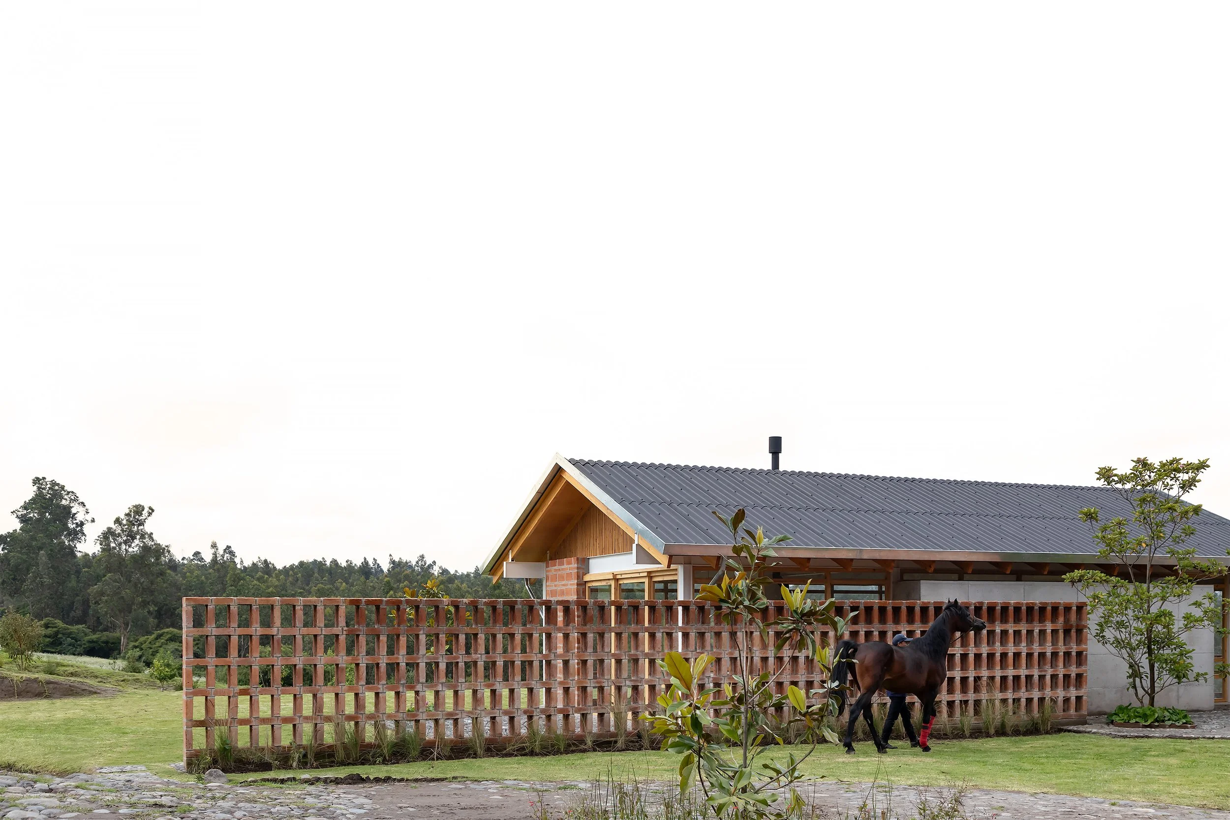 Casa de campo moderna con tejado de lámina metálica y muro de ladrillos, rodeada de un jardín con césped y árboles, en un entorno rural con montañas y árboles en el fondo, y un caballo negro en primer plano.