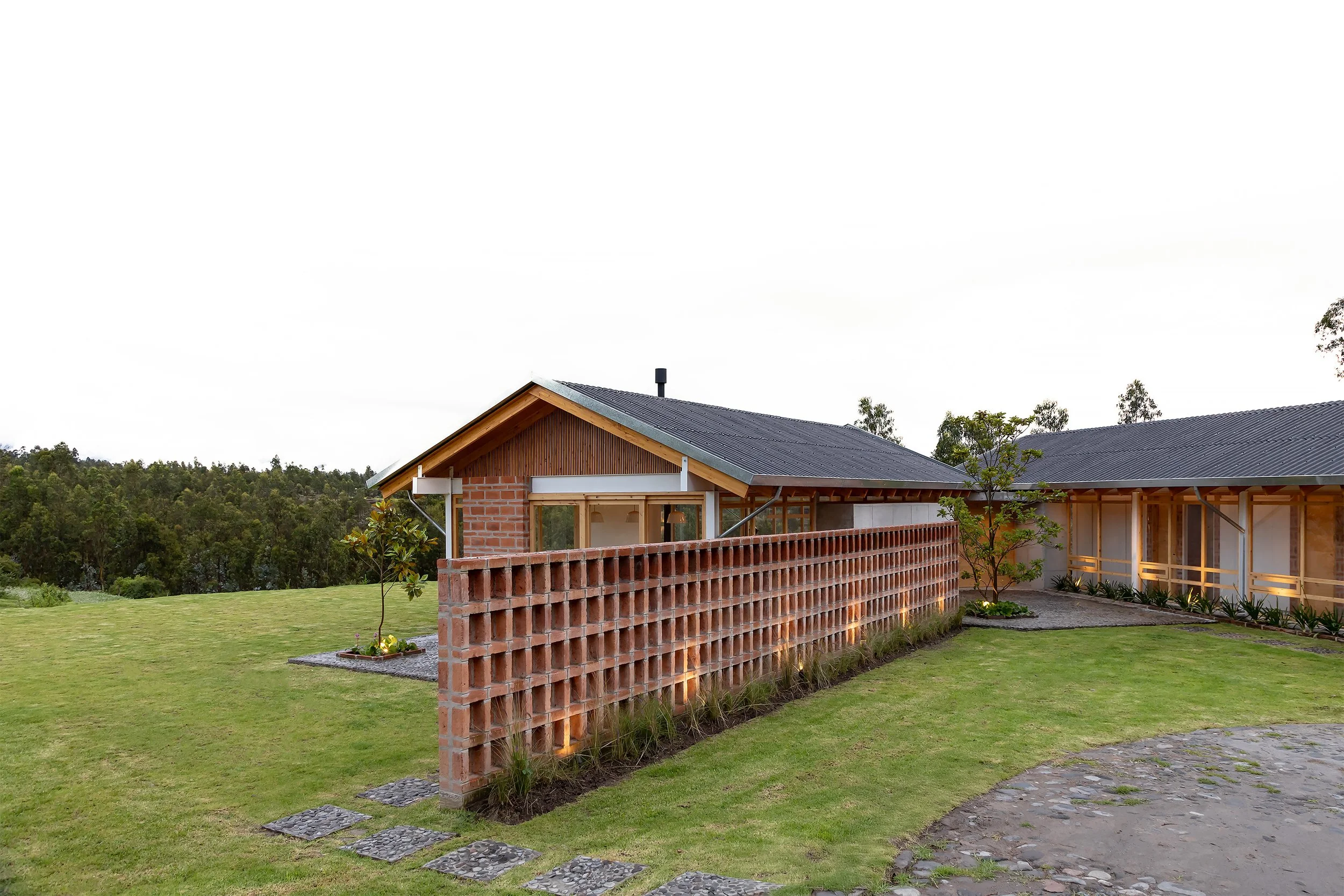 Casa en construcción con pared de bloques de arcilla, techos de lámina y ventanas de vidrio, rodeada de césped y árboles en un paisaje rural.