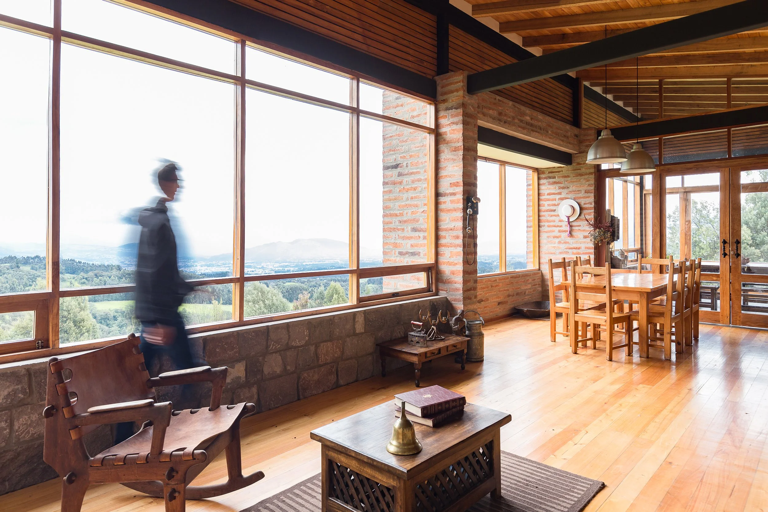 interior de una sala rústica con paredes de ladrillo, grandes ventanas y vista a la naturaleza, mesas y sillas de madera, y decoraciones sencillas.