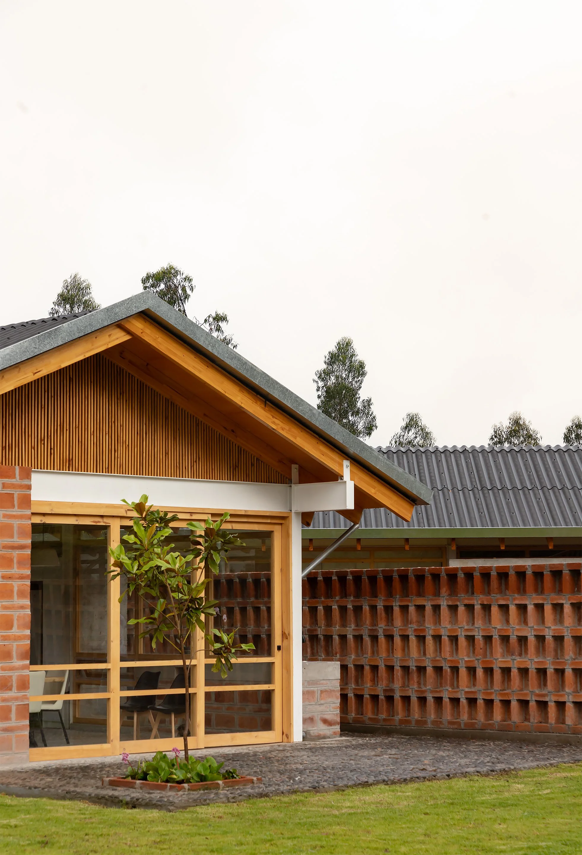 Casa moderna de ladrillo y madera con ventanas de vidrio, cerca de un césped y plantas, con un tejado de metal y árboles en el fondo, en día nublado.