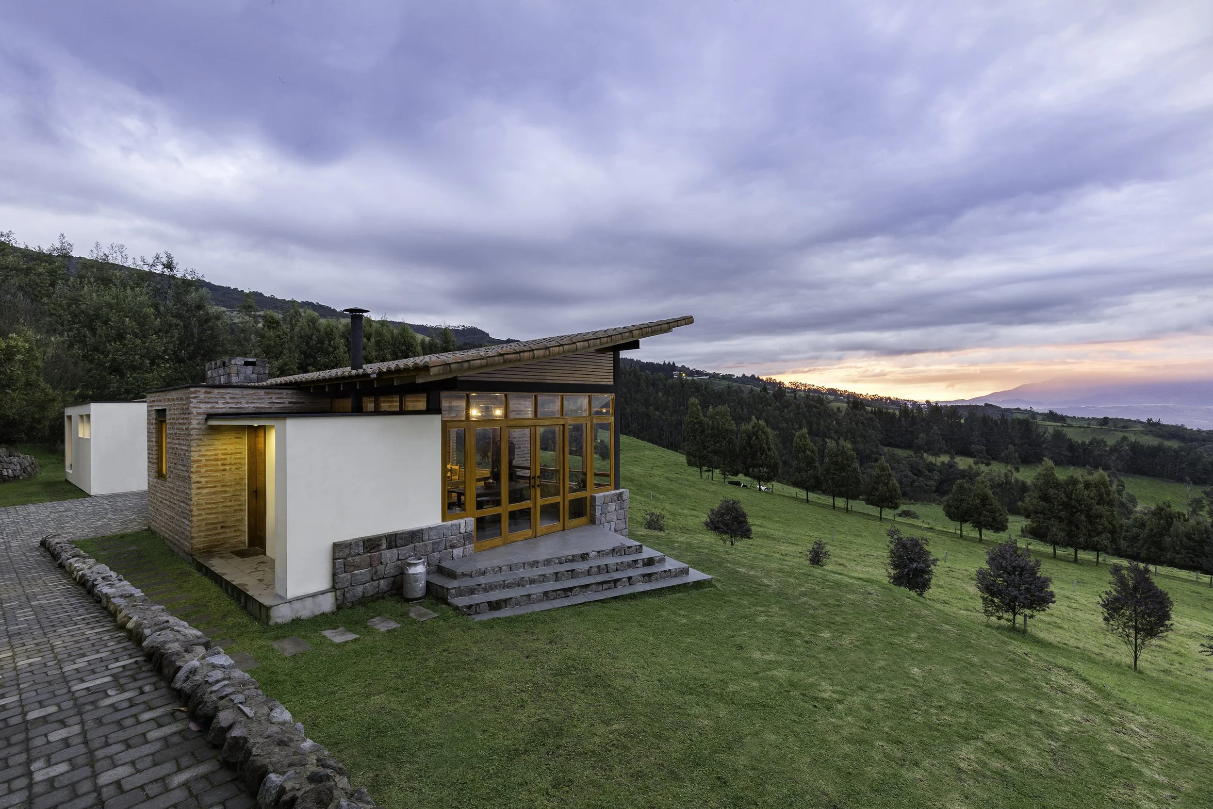 Casa moderna con grandes ventanas de madera, en un entorno rural con colinas verdes y árboles, al atardecer con cielo nublado.