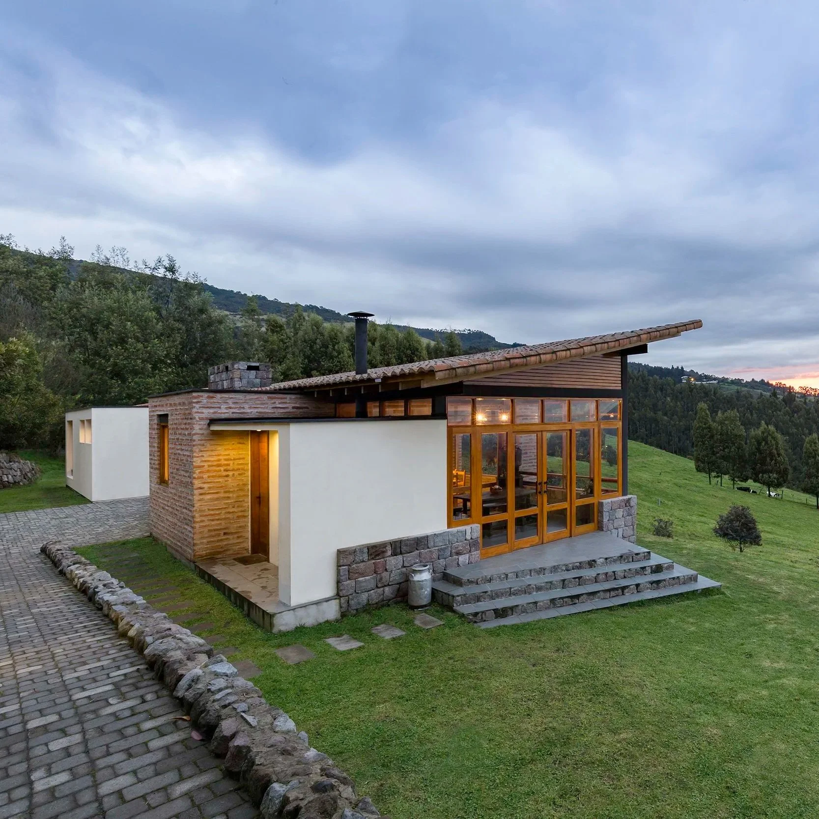 Casa moderna con ventanas de vidrio y techo inclinado, rodeada de césped y montañas al fondo, en un entorno natural.