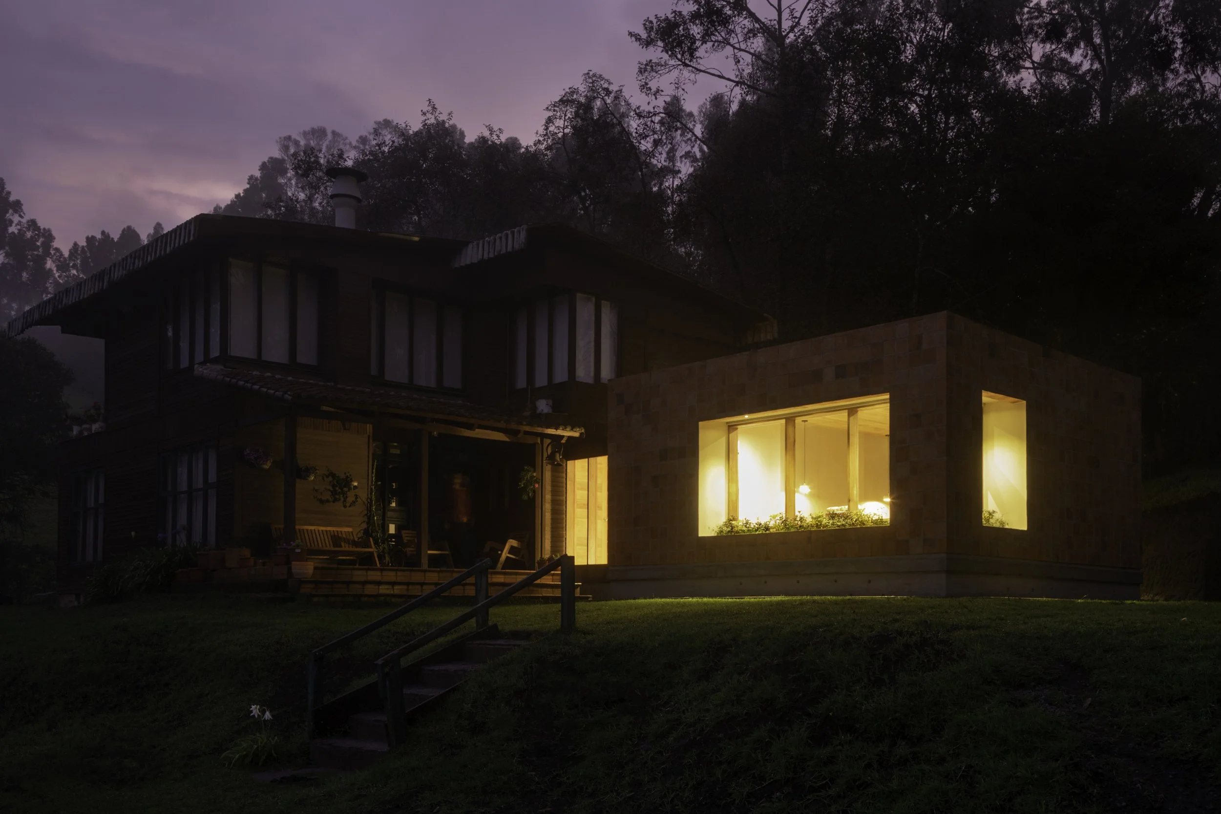 Casa moderna de dos pisos en la noche con ventanas iluminadas, rodeada de árboles y césped, cielo azul morado de atardecer o amanecer.