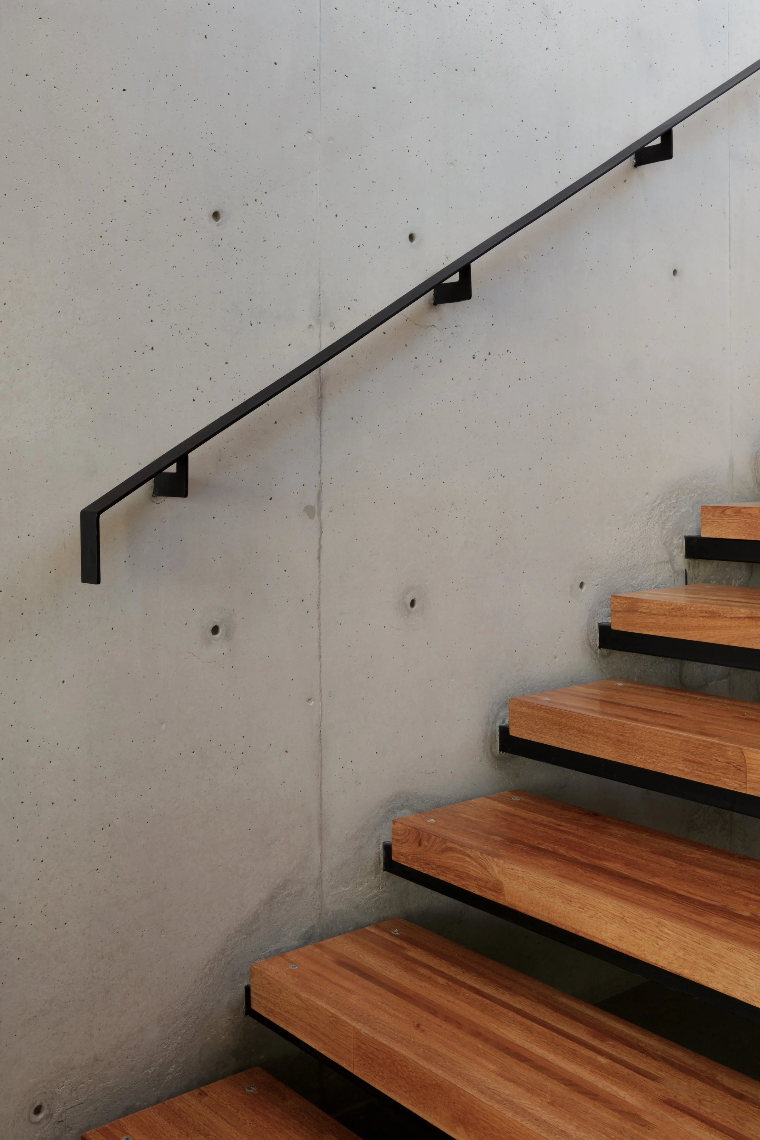 Escalera de madera con barandilla negra adhering a una pared de concreto de color gris claro con pequeñas perforaciones.