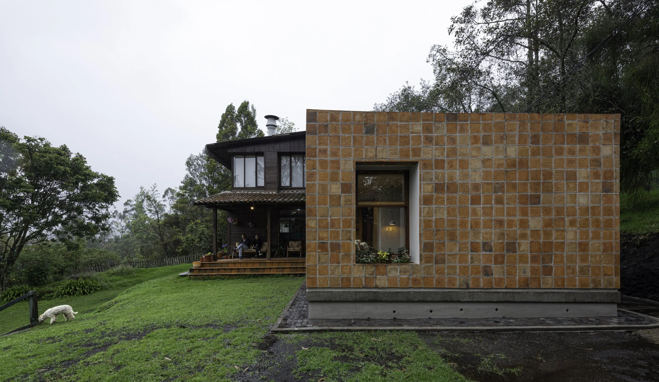 Casa moderna con fachada de ladrillos, ventana grande y porche con dos personas sentadas en sillones, rodeada de vegetación y árboles