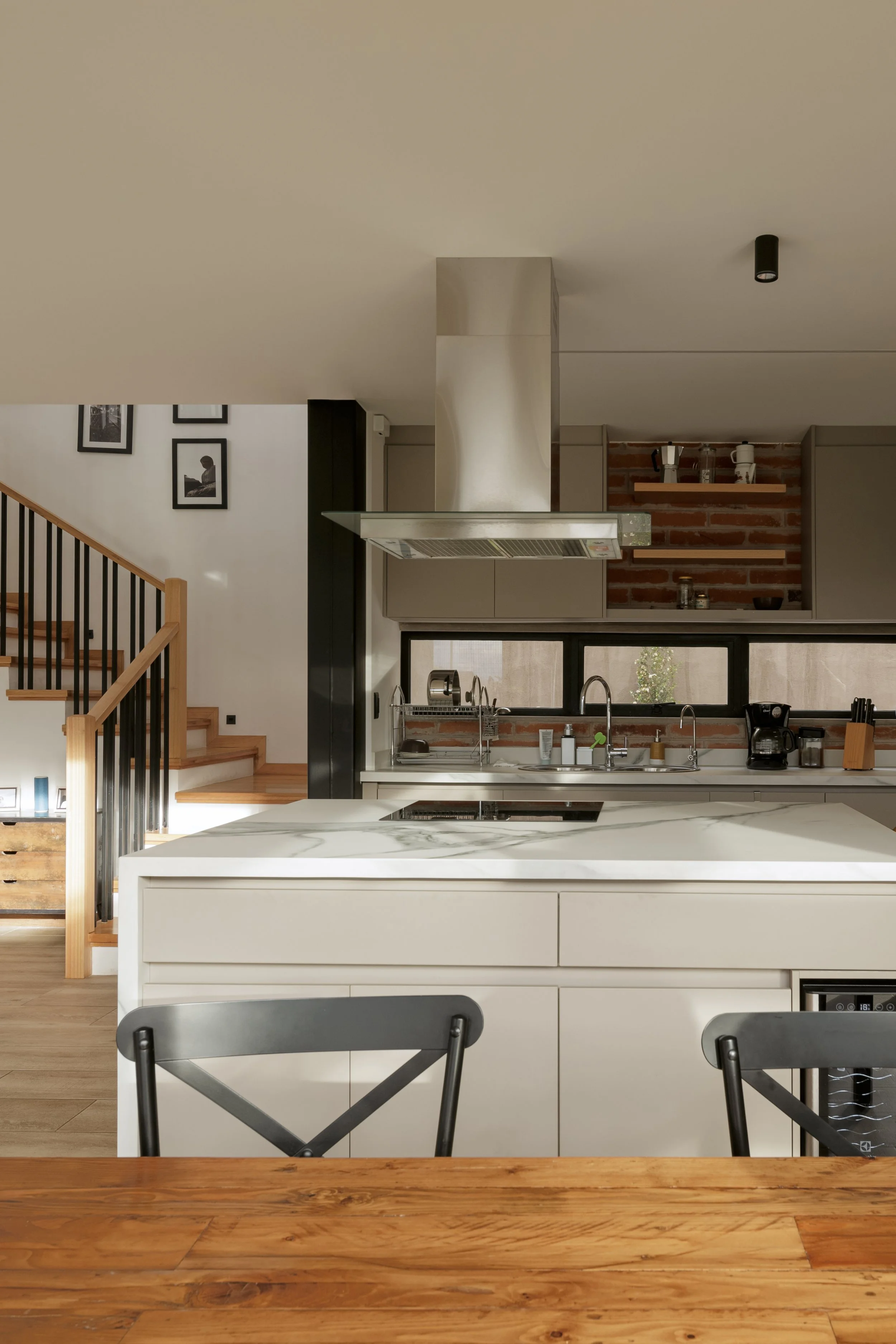 Cocina moderna con isla de mármol, estanterías de madera en muro de ladrillos y detalles en negro, escaleras de madera a la izquierda, paredes blancas y ventanas grandes.