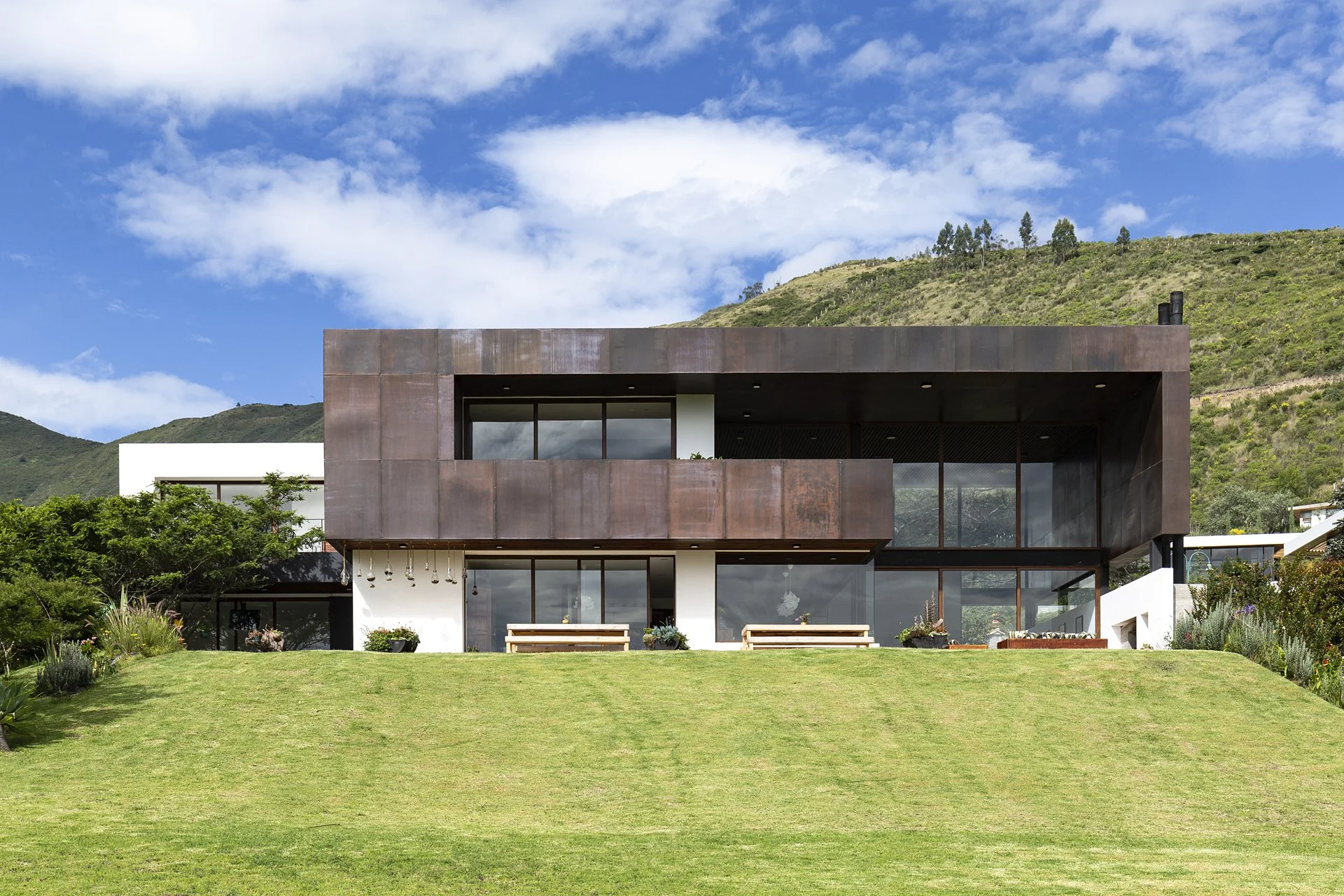 Casa moderna de dos pisos con paredes de metal y ventanas grandes, rodeada de un jardín verde bien cuidado, con colinas y cielo azul con algunas nubes al fondo.