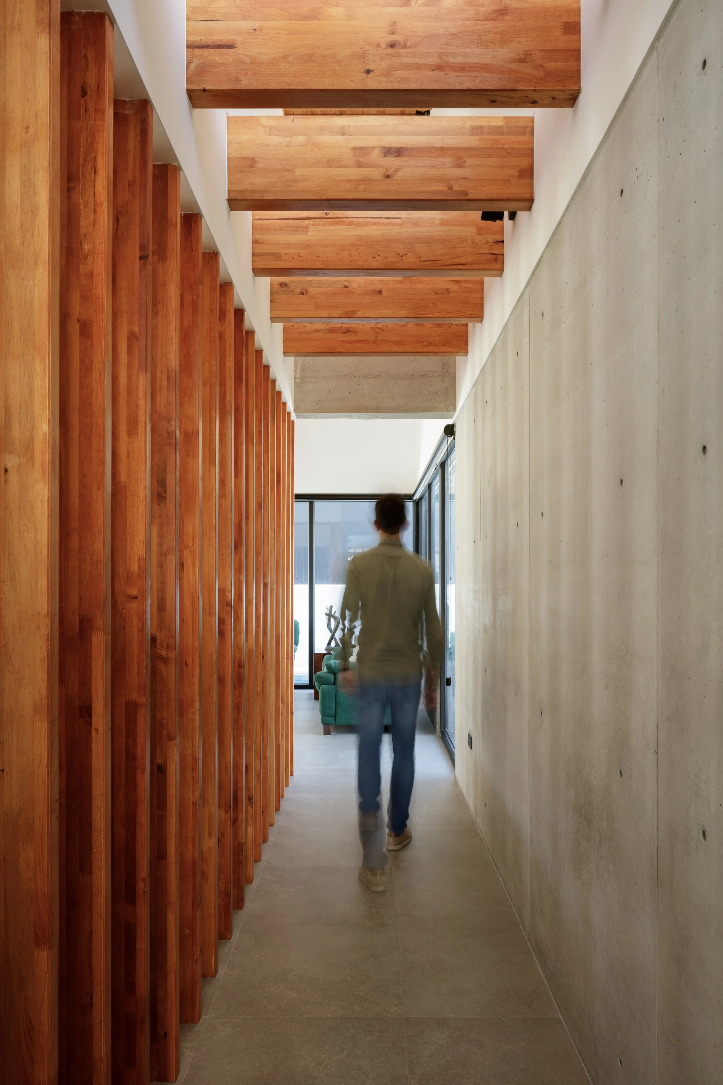 Un hombre camina por un pasillo interior con paredes de cemento a la derecha y paneles de madera a la izquierda, con un ventanal al fondo y una sala de estar visible a través de la ventana.