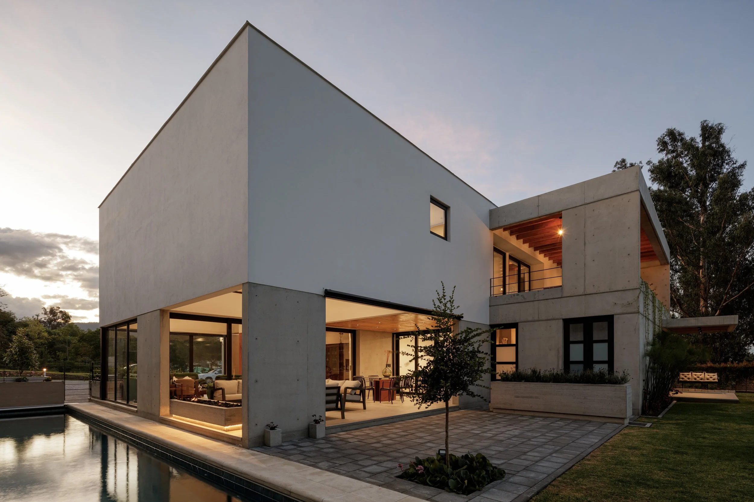 Casa moderna con diseño minimalista, ventanas grandes y área de patio con piscina, rodeada de árboles y bajo cielo despejado al atardecer.