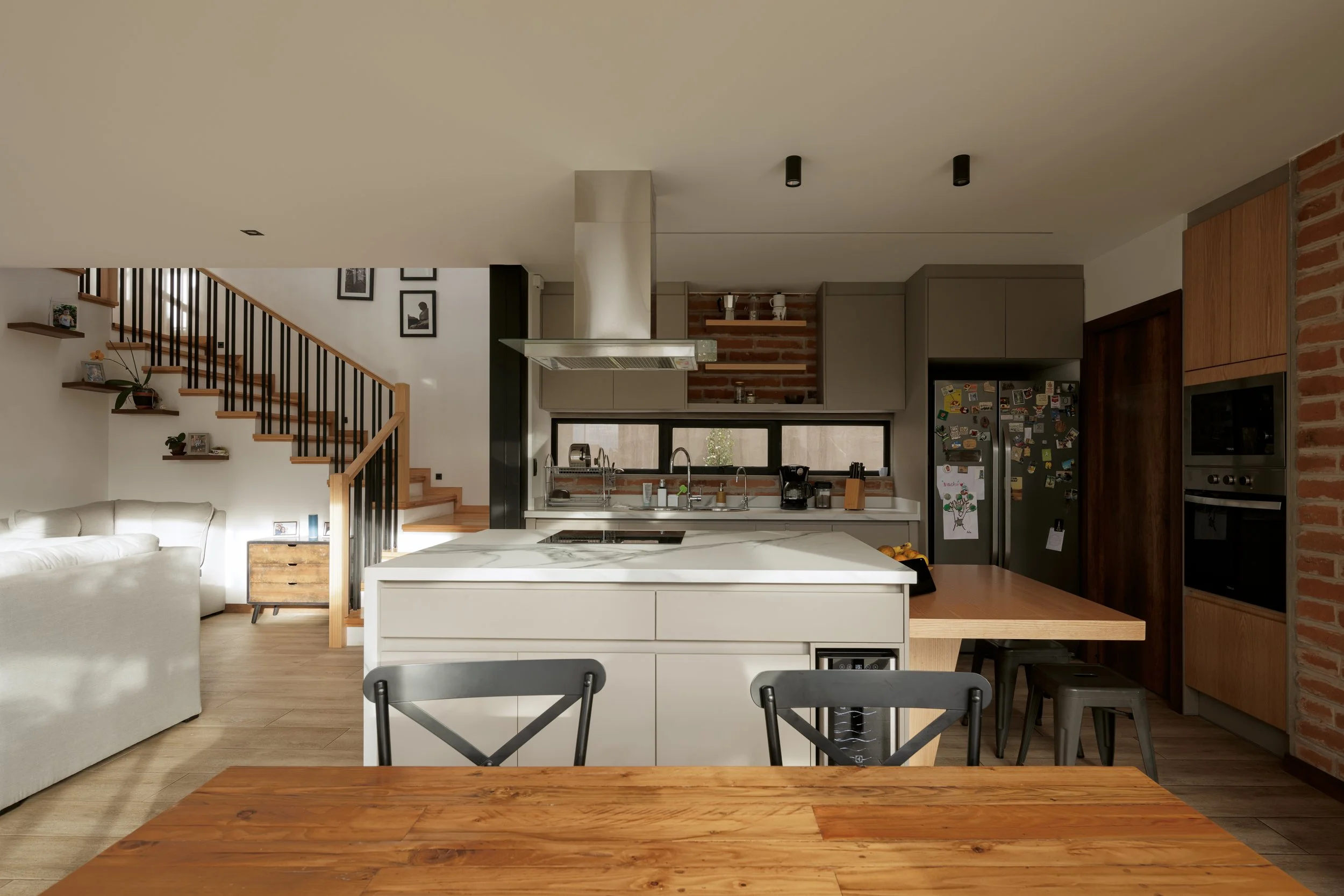 Vista de una cocina moderna con una isla blanca, electrodomésticos, estanterías y paredes de ladrillo, y una sala con espacio para comer.