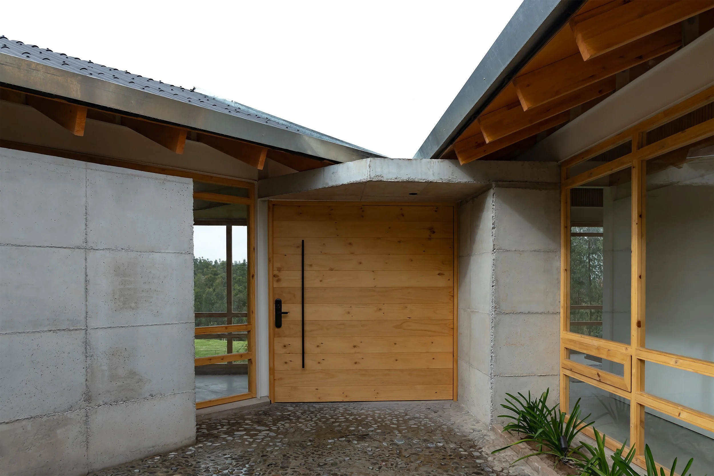 Entrada de una casa moderna con paredes de concreto, ventanas grandes con marcos de madera y puertas de madera, con techo inclinado de metal y estructura de madera visible, rodeada de naturaleza.