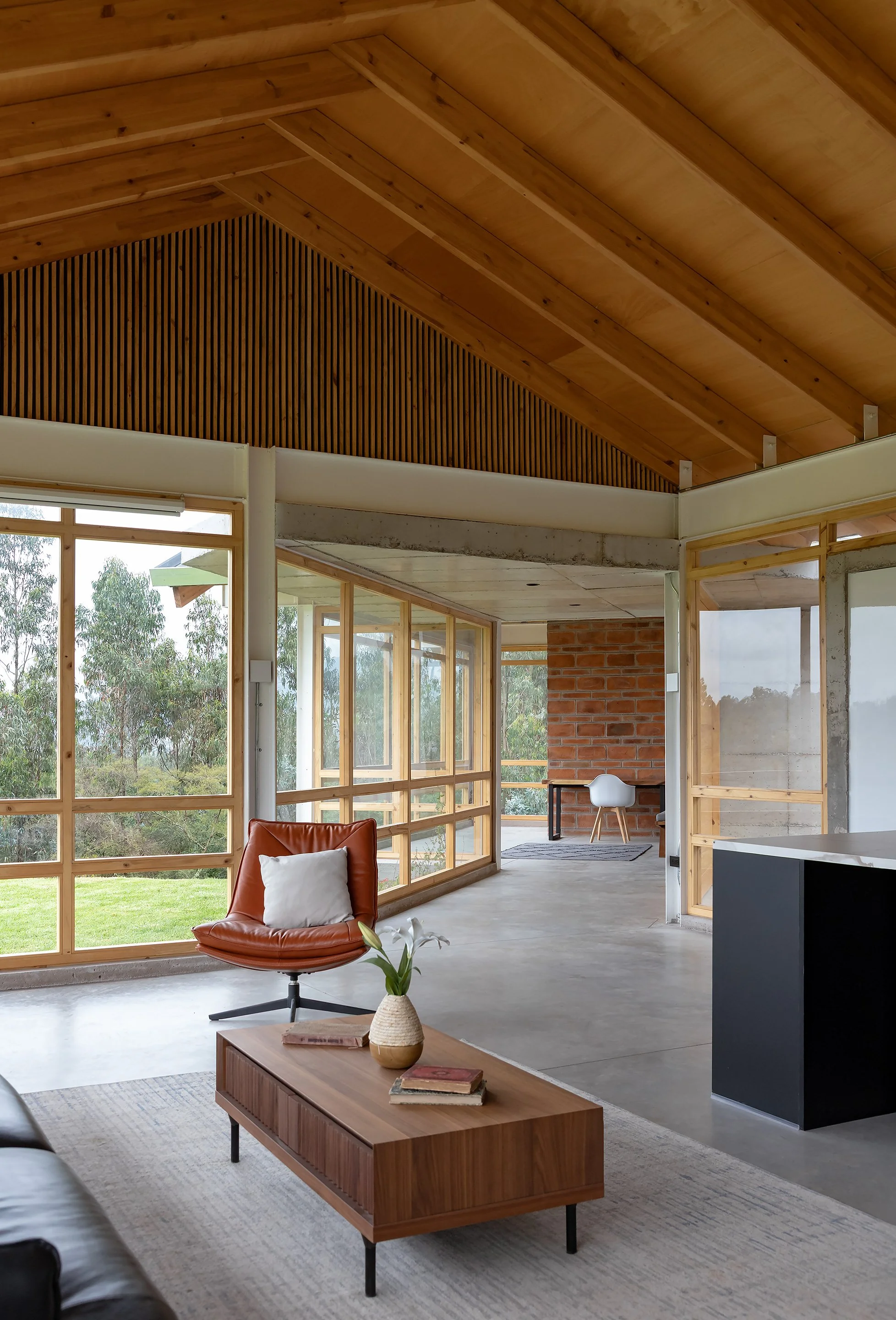 Interior de una sala moderna con paredes de madera, grandes ventanas de vidrio y muebles de diseño minimalista, incluyendo una silla de cuero color marrón, una mesa de centro de madera y una planta decorativa.