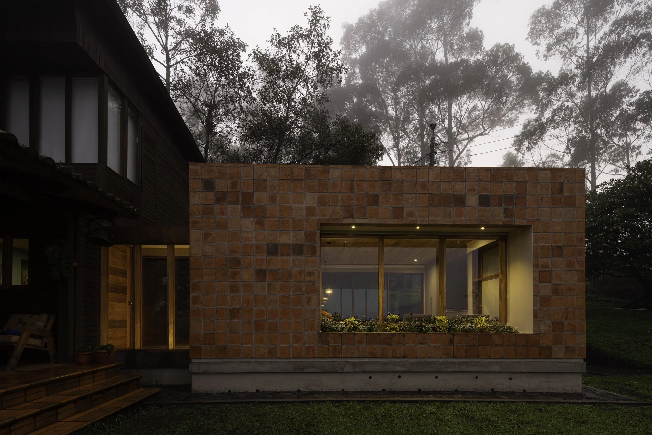 Casa moderna de ladrillos con ventanas grandes y un área de jardín, rodeada de árboles en un entorno neblinoso.