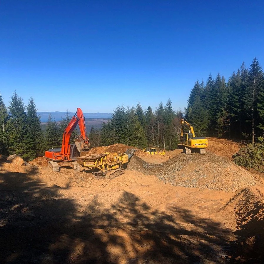 Construction site with excavators and gravel in a forested area under blue sky.