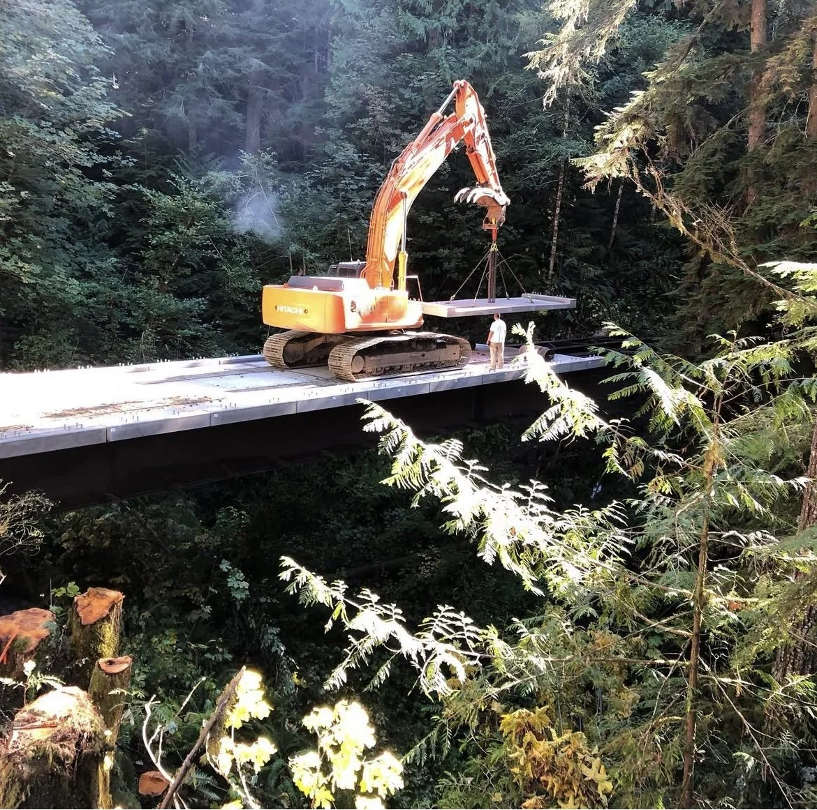 excavator placing a prefabricated bridge section on a bridge over a forested area.