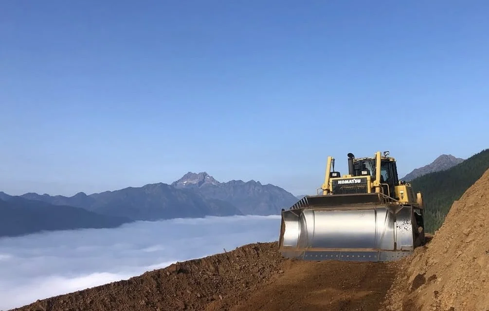 A bulldozer smoothing a dirt road on a mountain, with a fog-covered valley and distant mountains in the background during daytime.
