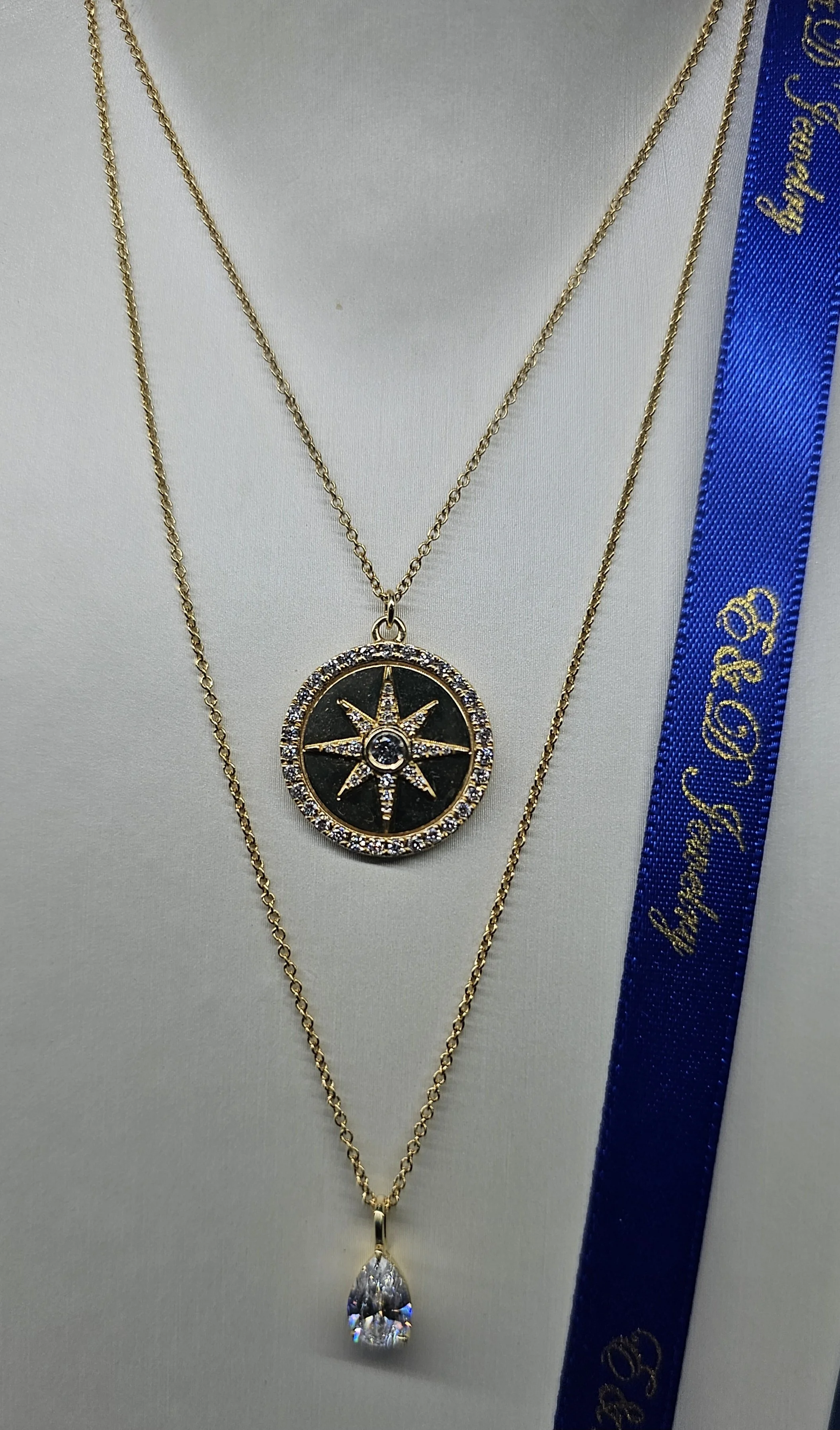 14 KT yellow gold layered necklaces with pendant, one featuring a compass design with rhinestones, and the other a large teardrop-shaped lab grown pear shaped diamond, displayed on a white neck holder with part of a blue ribbon with golden inprint.