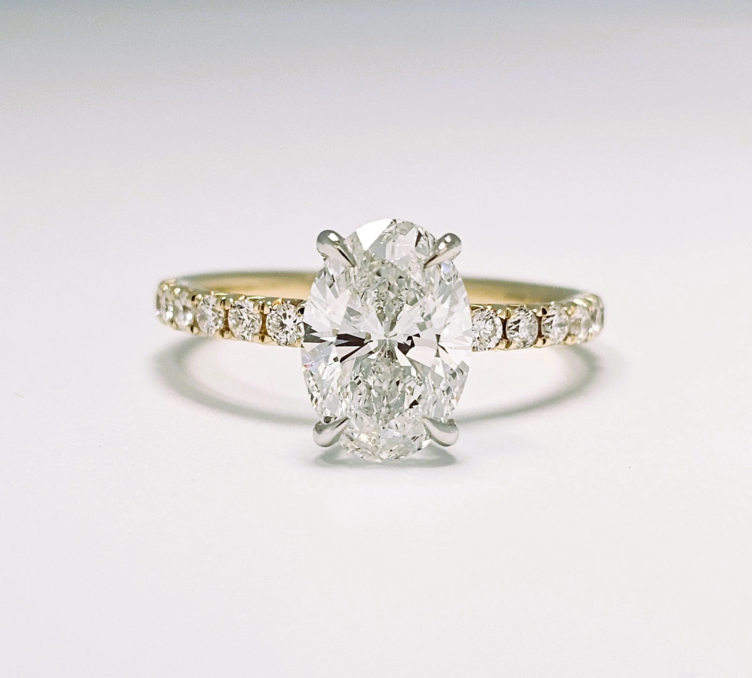 Close-up of a 14 kt yellow gold engagement ring with a large oval-shaped 3 karat diamond and smaller diamonds on the band, reflected on a shiny surface.