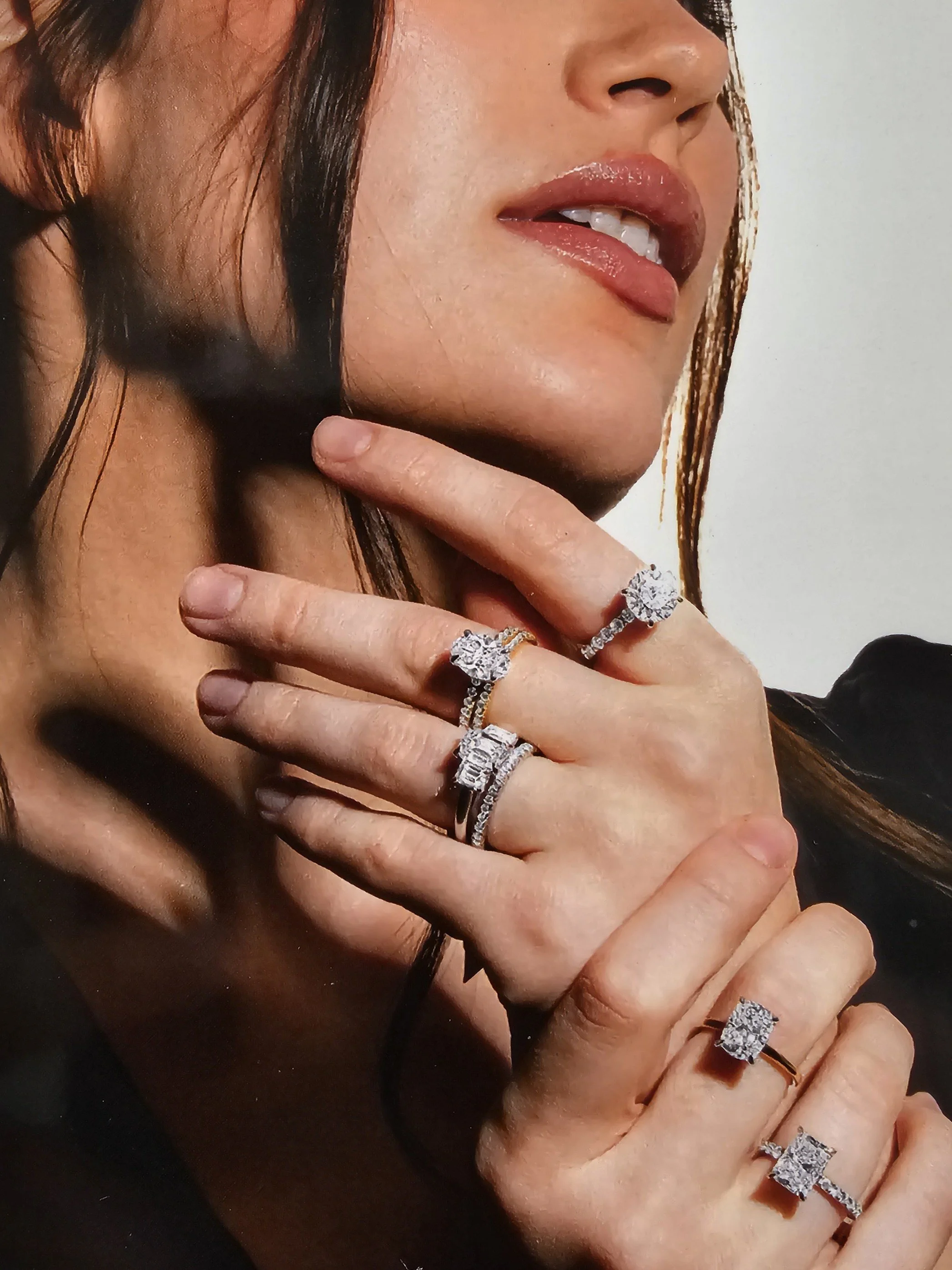 Close-up of a woman with long dark hair wearing multiple diamond rings on her fingers, touching her face with slightly parted lips.
