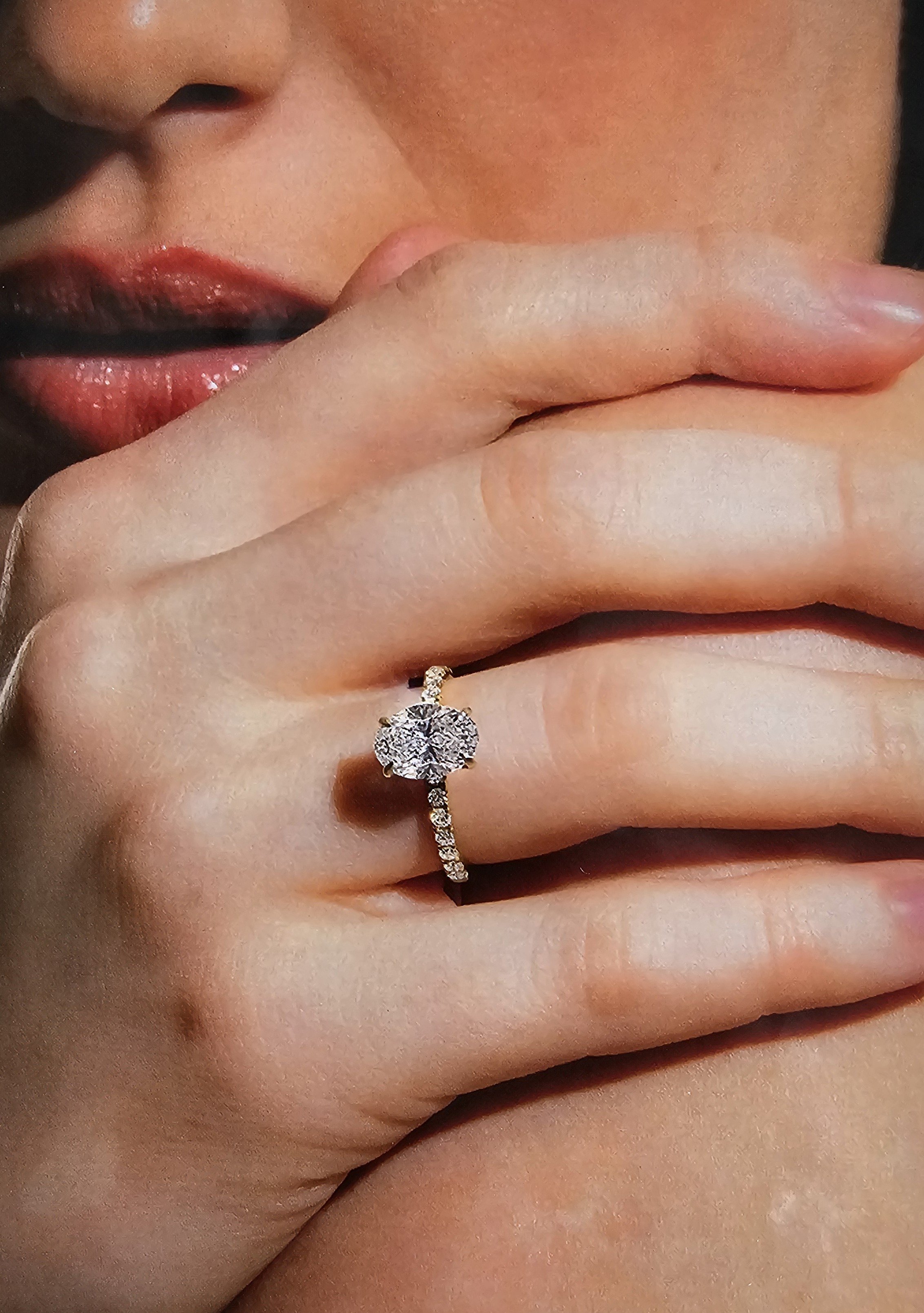 Close-up of a person's face and hand, with a focus on lips, finger, and diamond ring with a large round center stone surrounded by smaller diamonds.