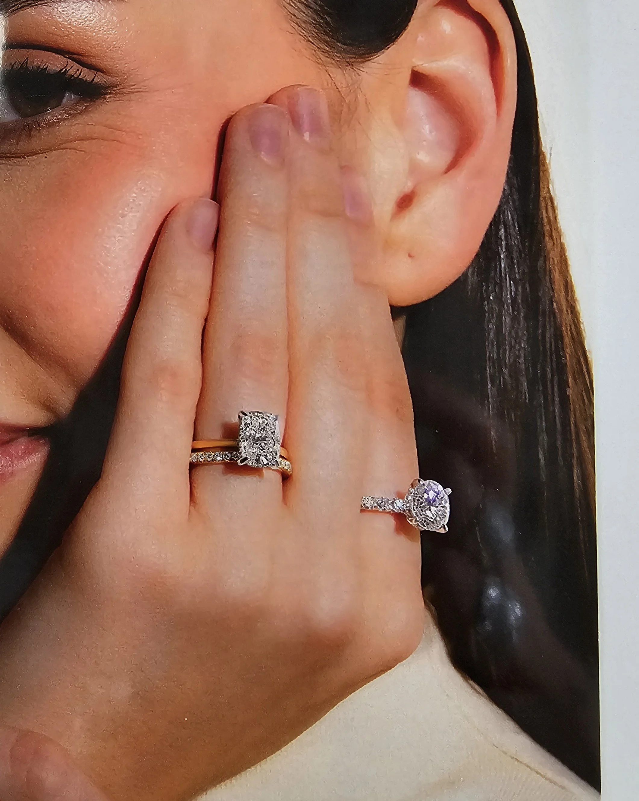 Close-up of a woman with shiny skin, black hair, and eye makeup, holding her fingers over her nose, revealing rings and rings with large stones.