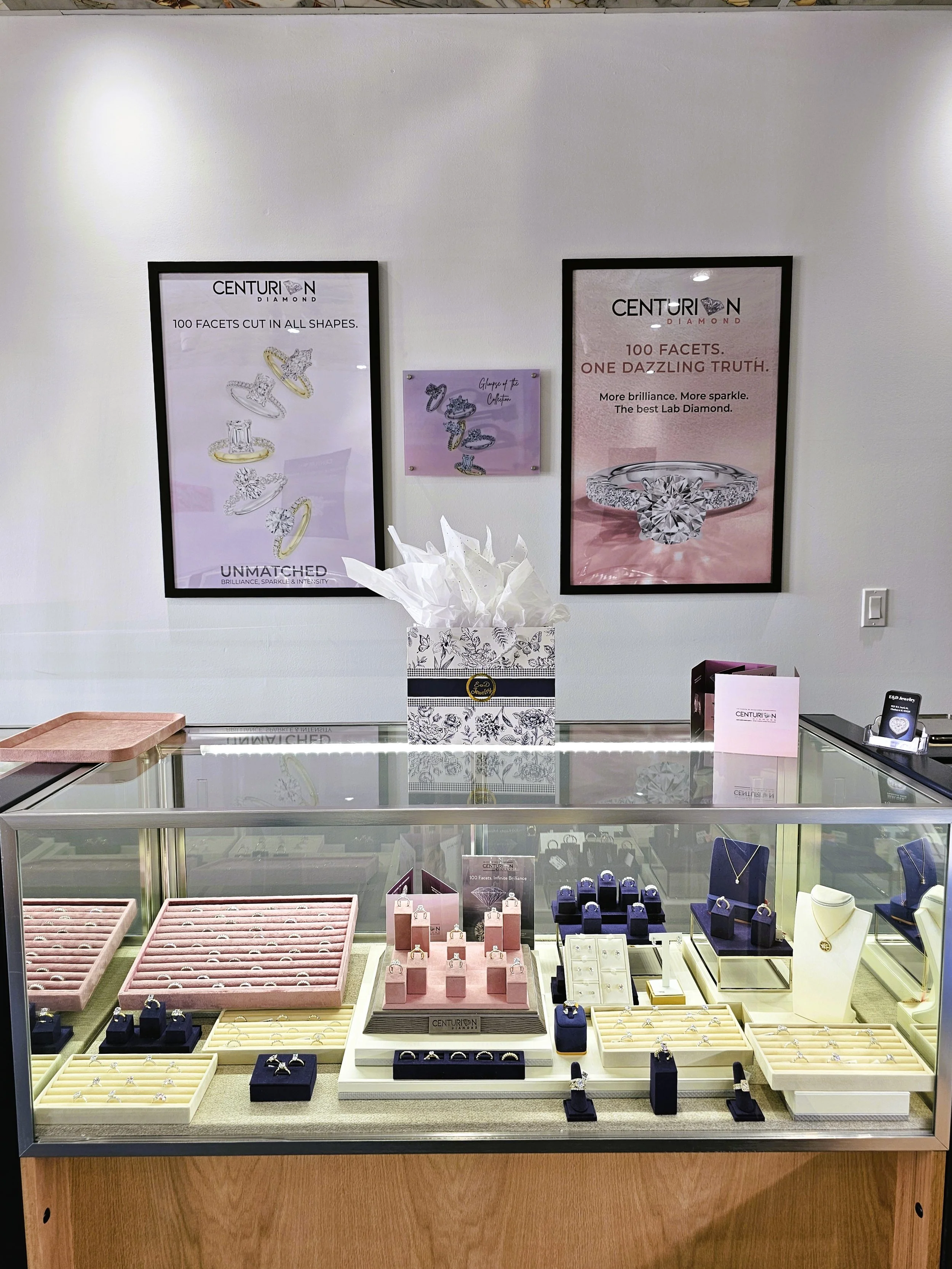 A jewelry display case with various rings, necklaces, and earrings, wi on the wall advertising diamond jewelry, including a large diamond ring highlighting 100 facets and the tagline 'More brilliance. More sparkle. The best Lab Diamond.'