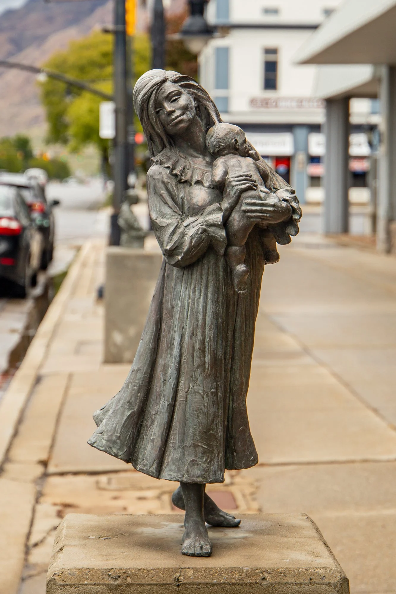 A bronze statue of a woman with long hair holding a child, standing on a concrete base on a sidewalk in a small town.