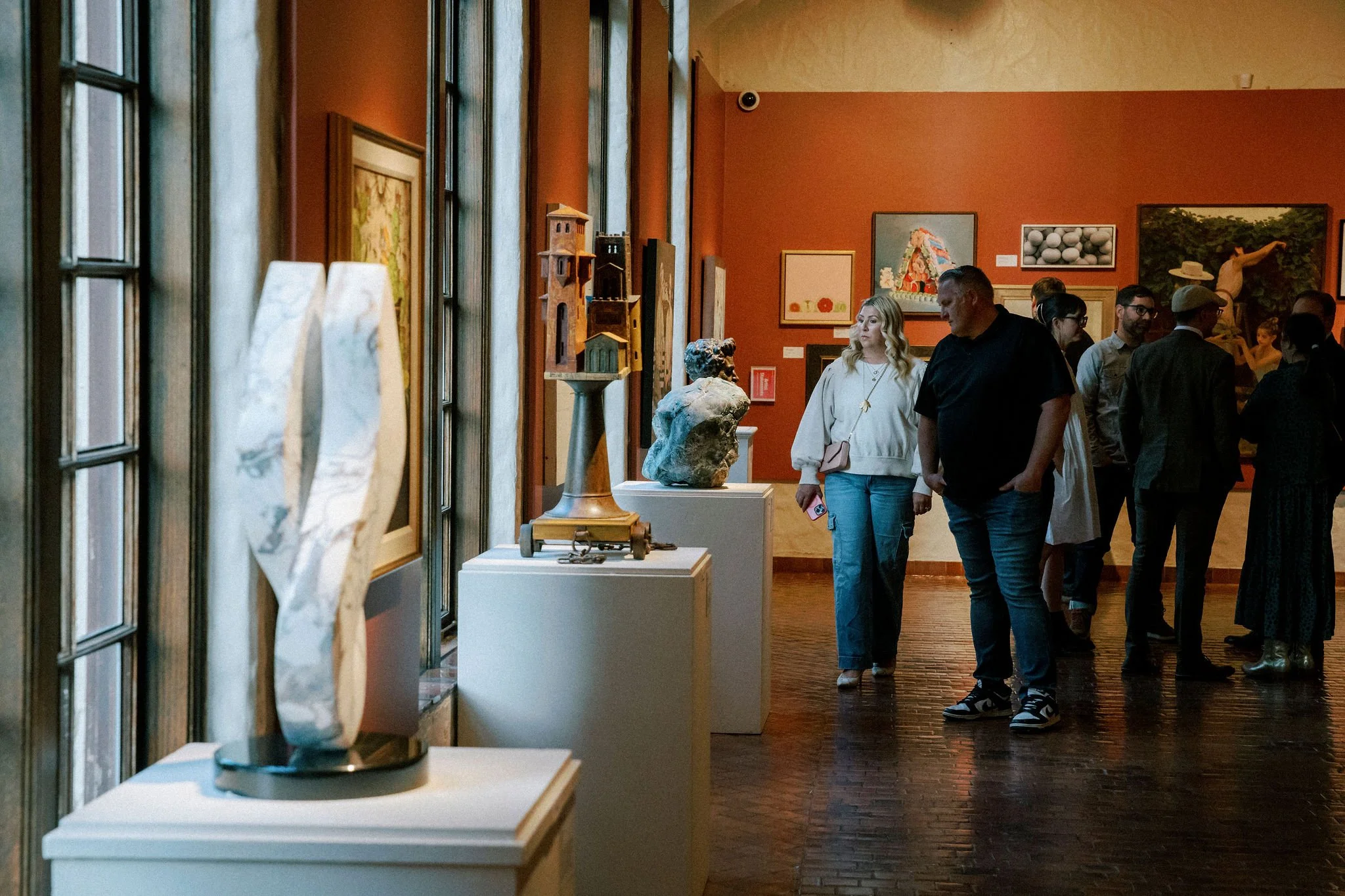 Visitors walking through an art gallery with paintings and sculptures on display, captured from a side angle near windows.