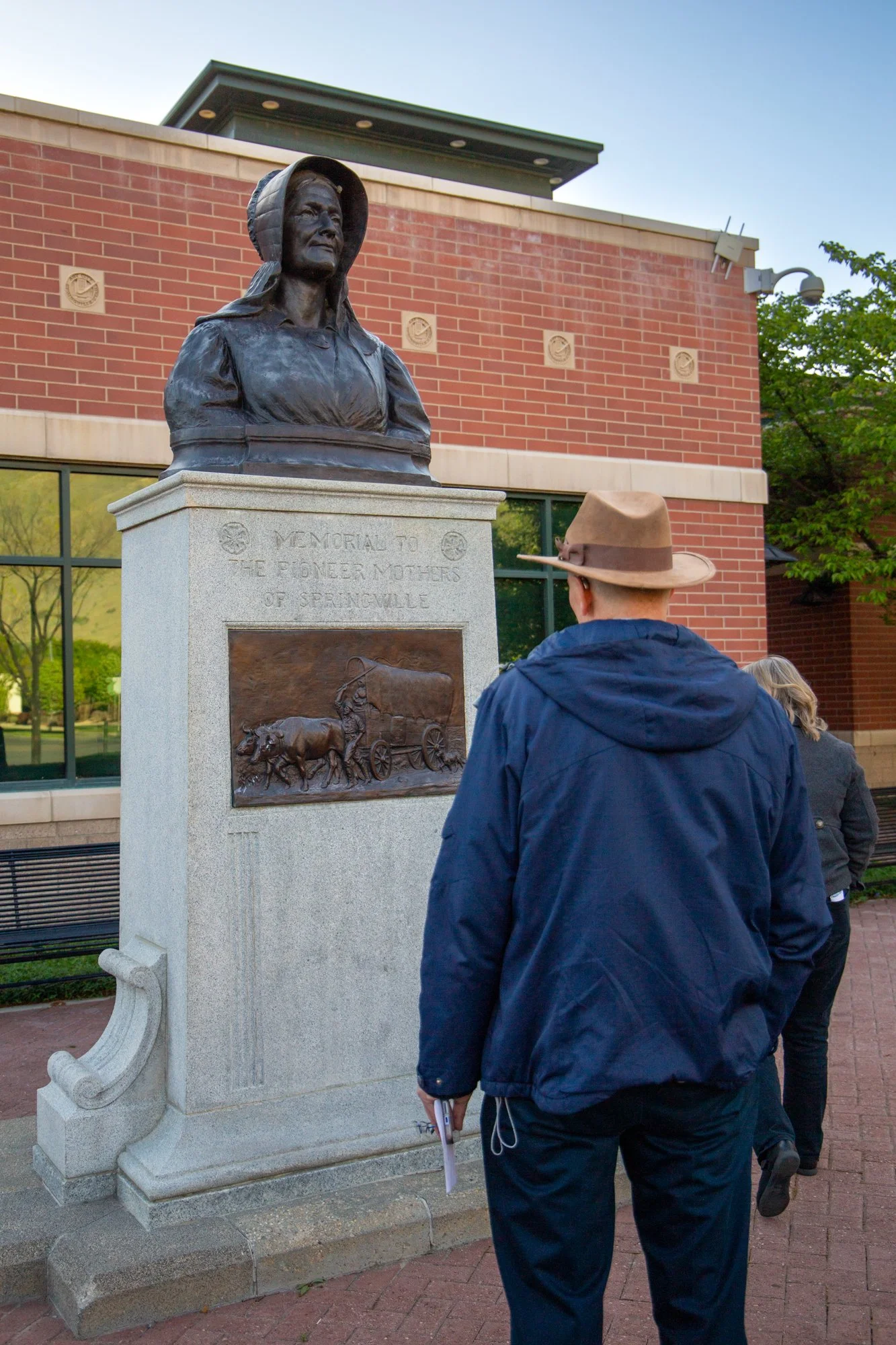 A person wearing a brown hat and navy jacket stands in front of a bronze bust monument of a woman with a hood, placed on a stone pedestal with an engraved plaque. The plaque remembers the Pioneer Mothers of Spokane.