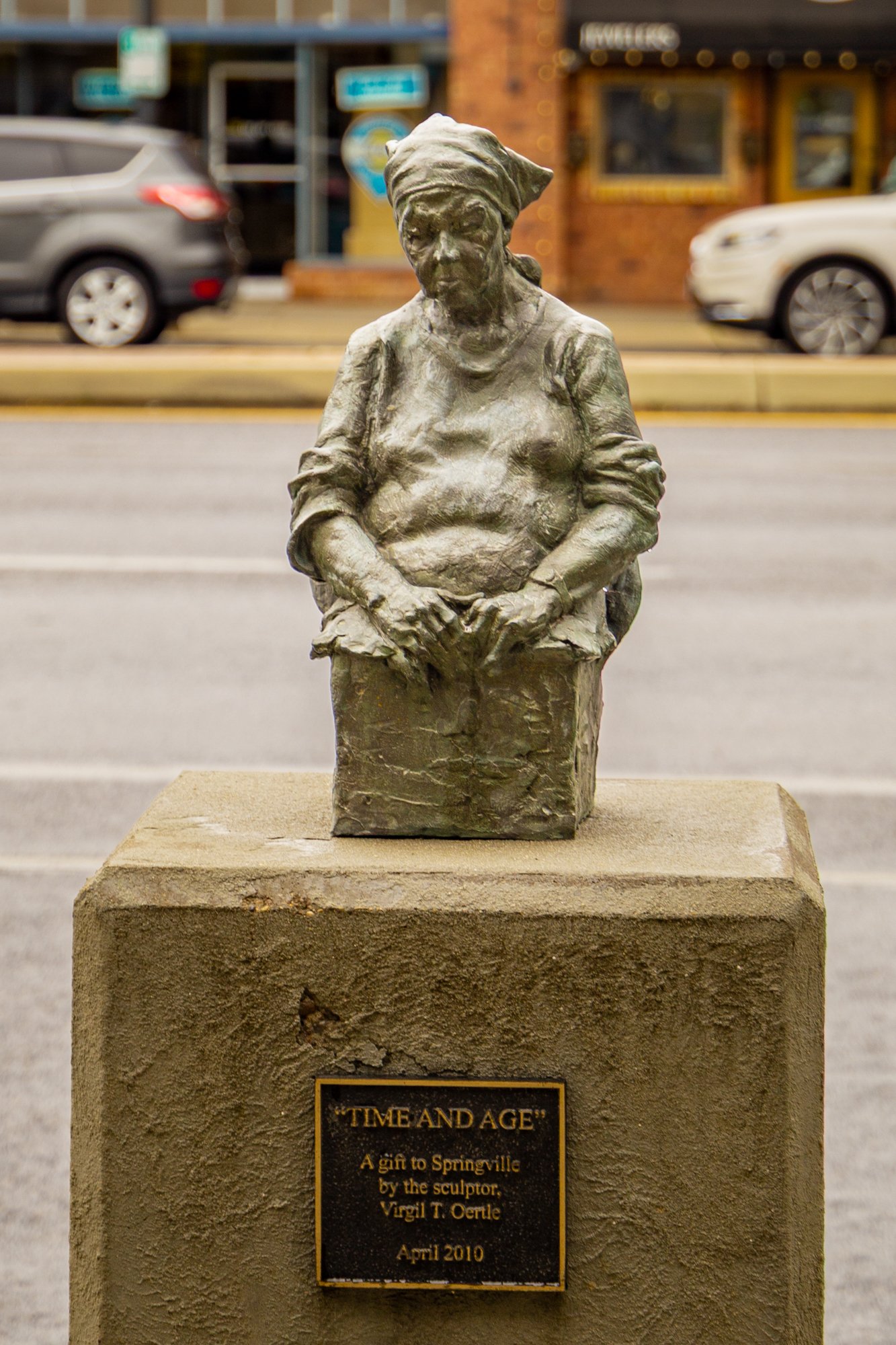 Bronze sculpture of a woman wearing a headscarf and casual clothes, standing with her hands clasped in front of her. The sculpture is on a stone pedestal with a plaque that reads: "TIME AND AGE, A gift to Springville by the sculptor Virgil T. Oertle,