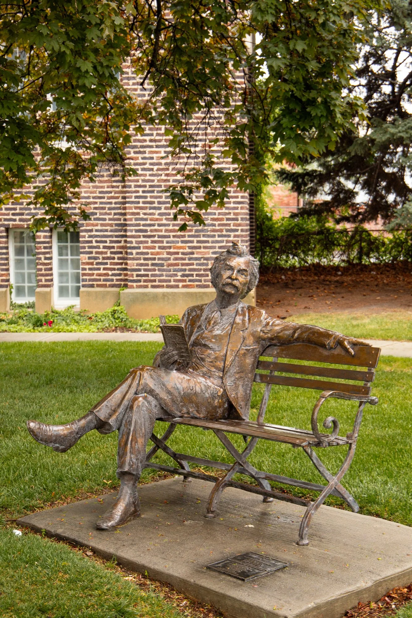 Bronze statue of a woman reading a book while reclining on a park bench in a garden area, surrounded by green grass and trees, with a brick building in the background.