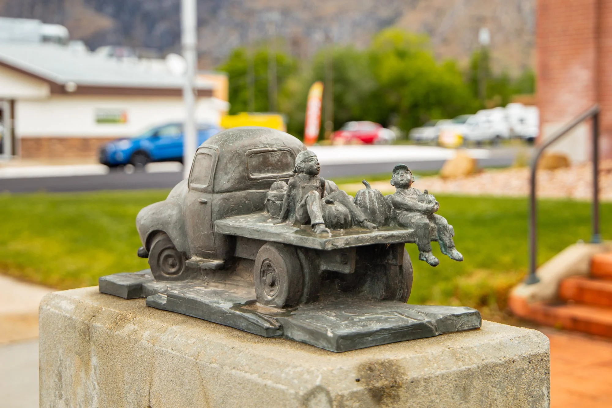 A bronze sculpture of two children riding with backpacks and pumpkins on a flatbed on a toy truck, set outdoors in a park with grass, trees, and a parking lot in the background.