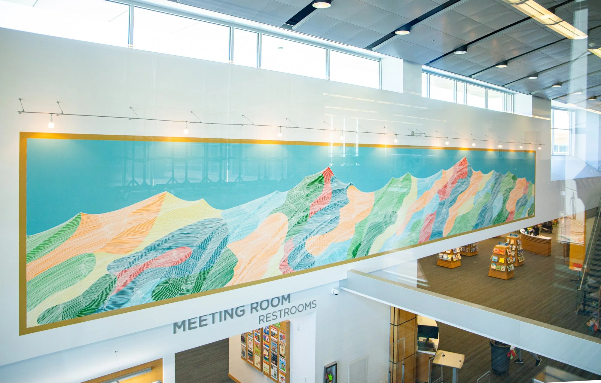 Interior of a modern building with large windows, showing a colorful abstract mural of mountain-like shapes with lines in various shades of orange, pink, green, blue, and yellow. Part of a bookstore or gift shop is visible below the mural.