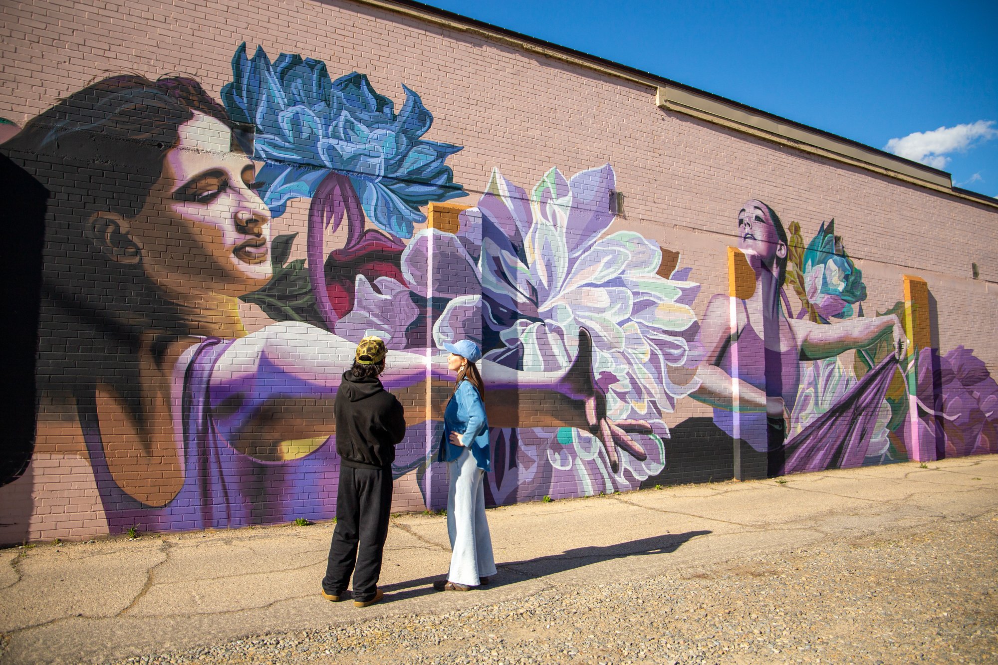 A mural painted on a brick wall depicting two women with large flowers, one with her hand extended. Two women are standing in front of the mural, talking, in bright daylight against a clear blue sky.