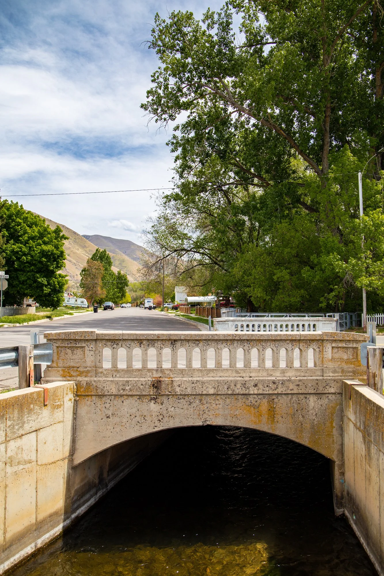 A small bridge over a dark water canal, with large green trees on the right and a street with parked cars and mountains in the background.