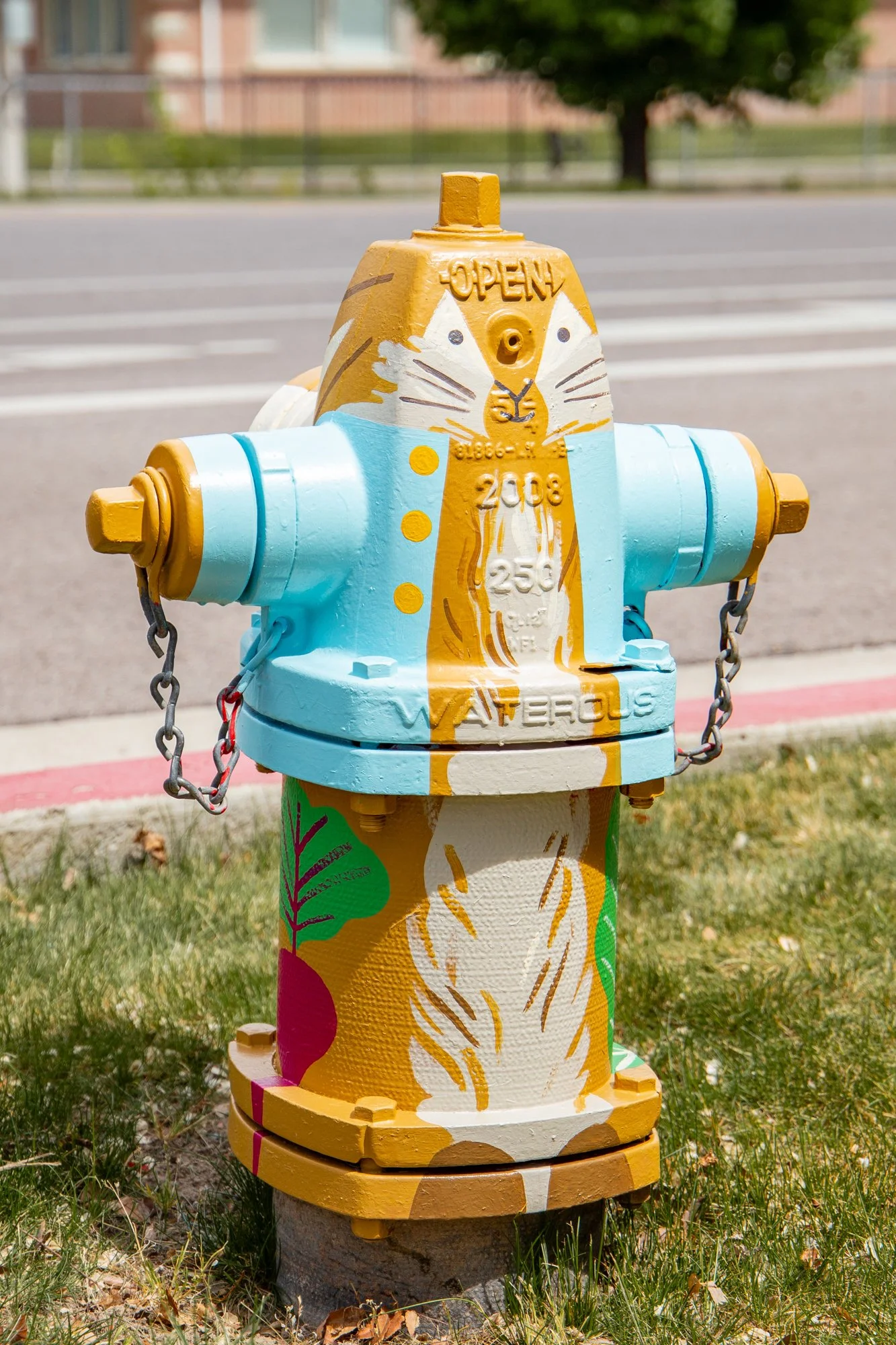 Fire hydrant decorated with colorful paint, featuring a dog face illustration with whiskers, ears, and an 'OPEN' sign on top, located in a grassy area with a sidewalk, trees, and a building in the background.