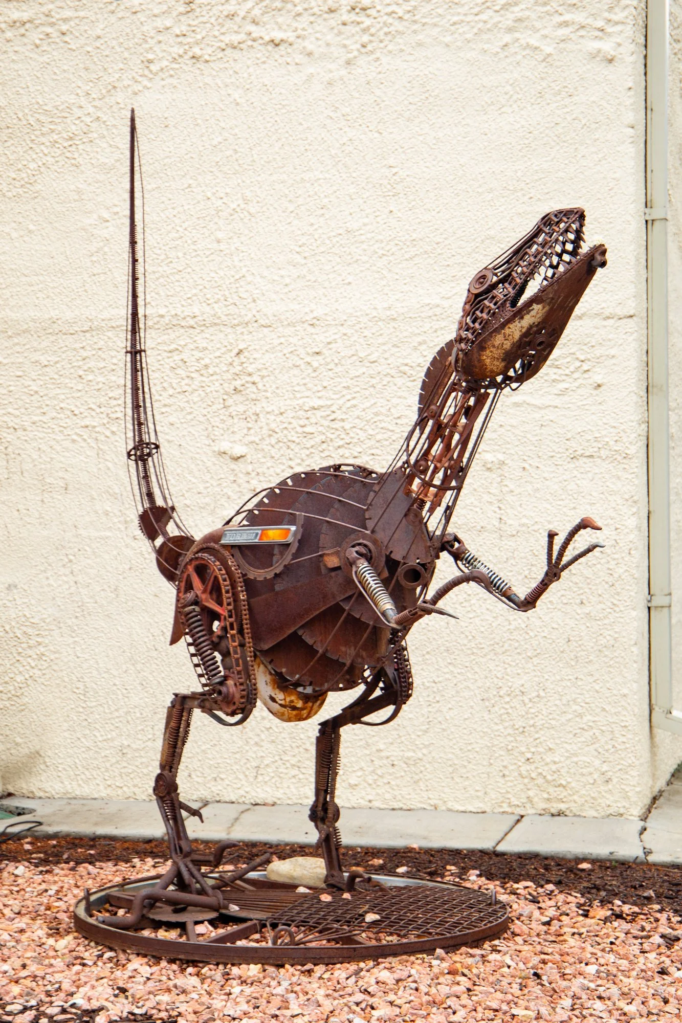Metal sculpture of a running horse made from rusted metal parts, standing on a circular metal base on an outdoor surface with small rocks.