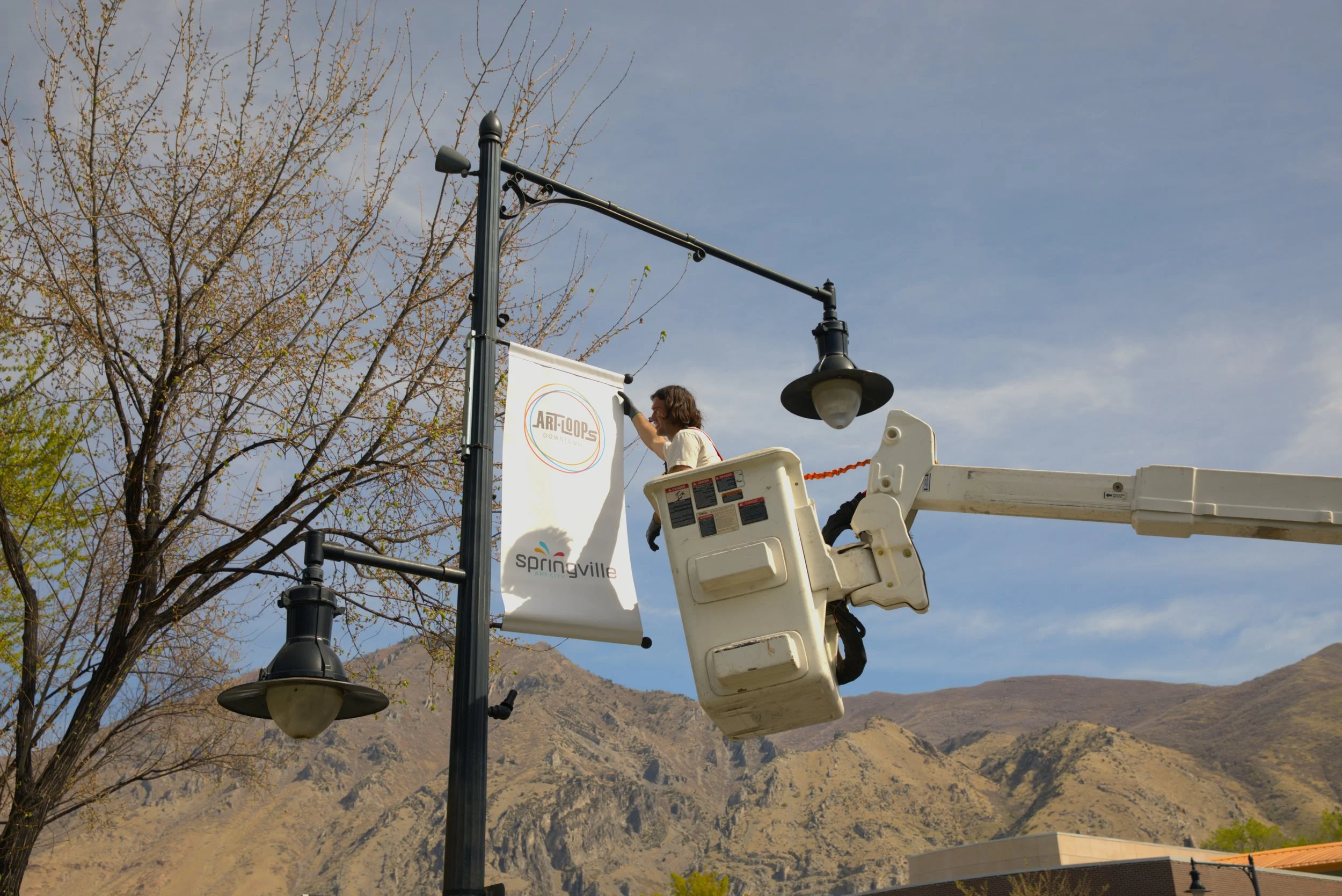 A person in a white shirt working on a streetlamp with a lift bucket. The streetlamp has a banner attached to it that reads 'ART LOOPS' and 'Springville.' There are leafless trees and mountains in the background.