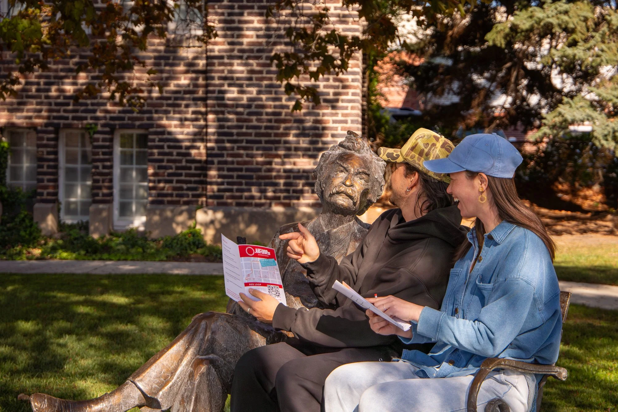 Two women sitting on a park bench and looking at a map together, next to a bronze bust statue of a man with a mustache, trees, and brick buildings in the background.