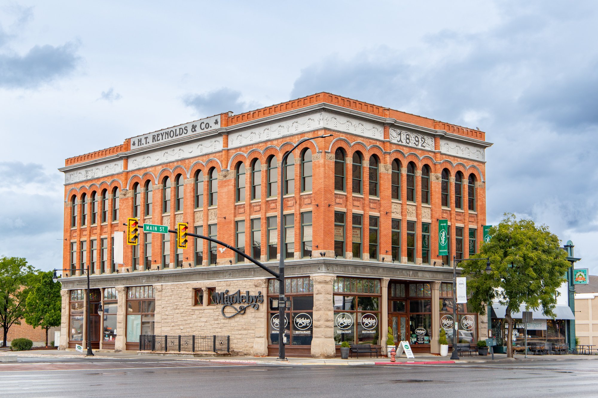 A historic red brick building with white decorative accents and large arched windows, situated on a city street corner with a sign reading Main St. and a restaurant named Mapleys at the ground level.