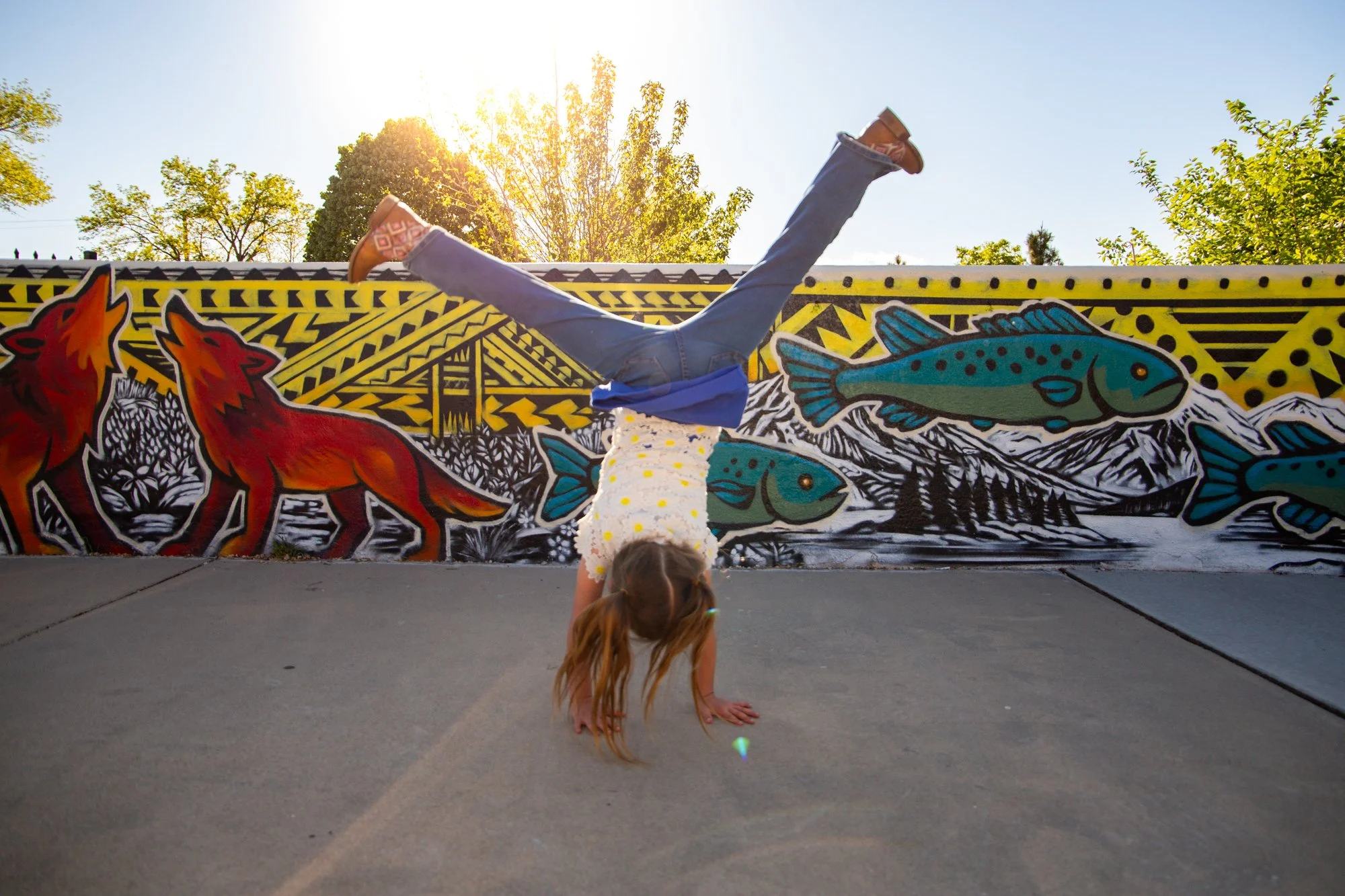 A young girl with long hair doing a handstand on the sidewalk in front of a colorful mural with wolves, fish, and mountains, with trees and a blue sky in the background.