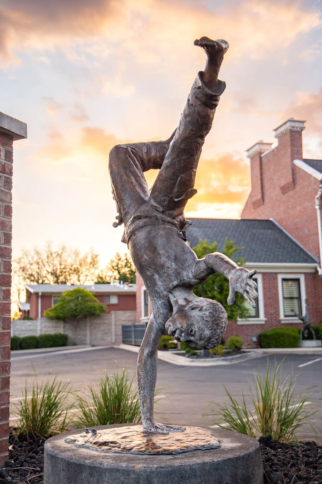 A bronze sculpture of a gymnast performing a handstand on a blog, with a sunset sky and residential buildings in the background.