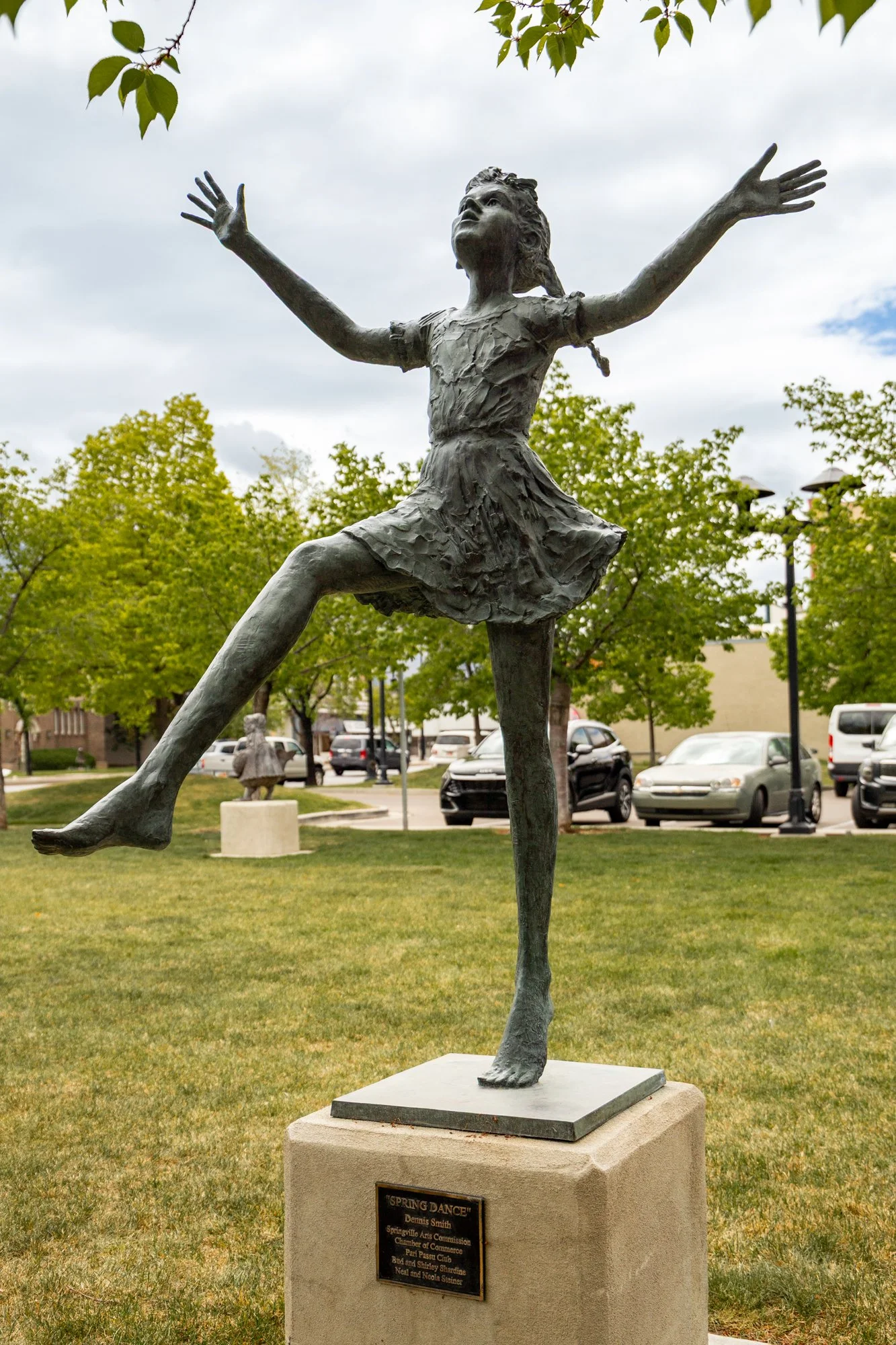 Bronze sculpture of a girl in a spring dance pose with arms outstretched and one leg raised, on a stone pedestal in a park with green trees, cars, and buildings in the background.