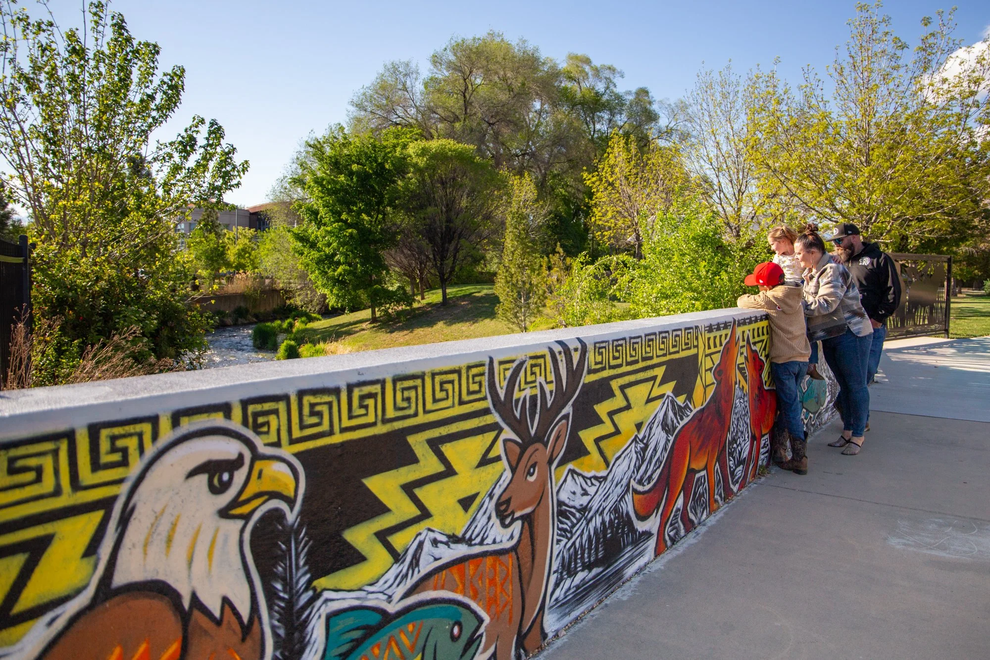 A group of four people leaning over a brightly painted mural on a low wall in an outdoor park with trees and a creek in the background, under a clear blue sky.