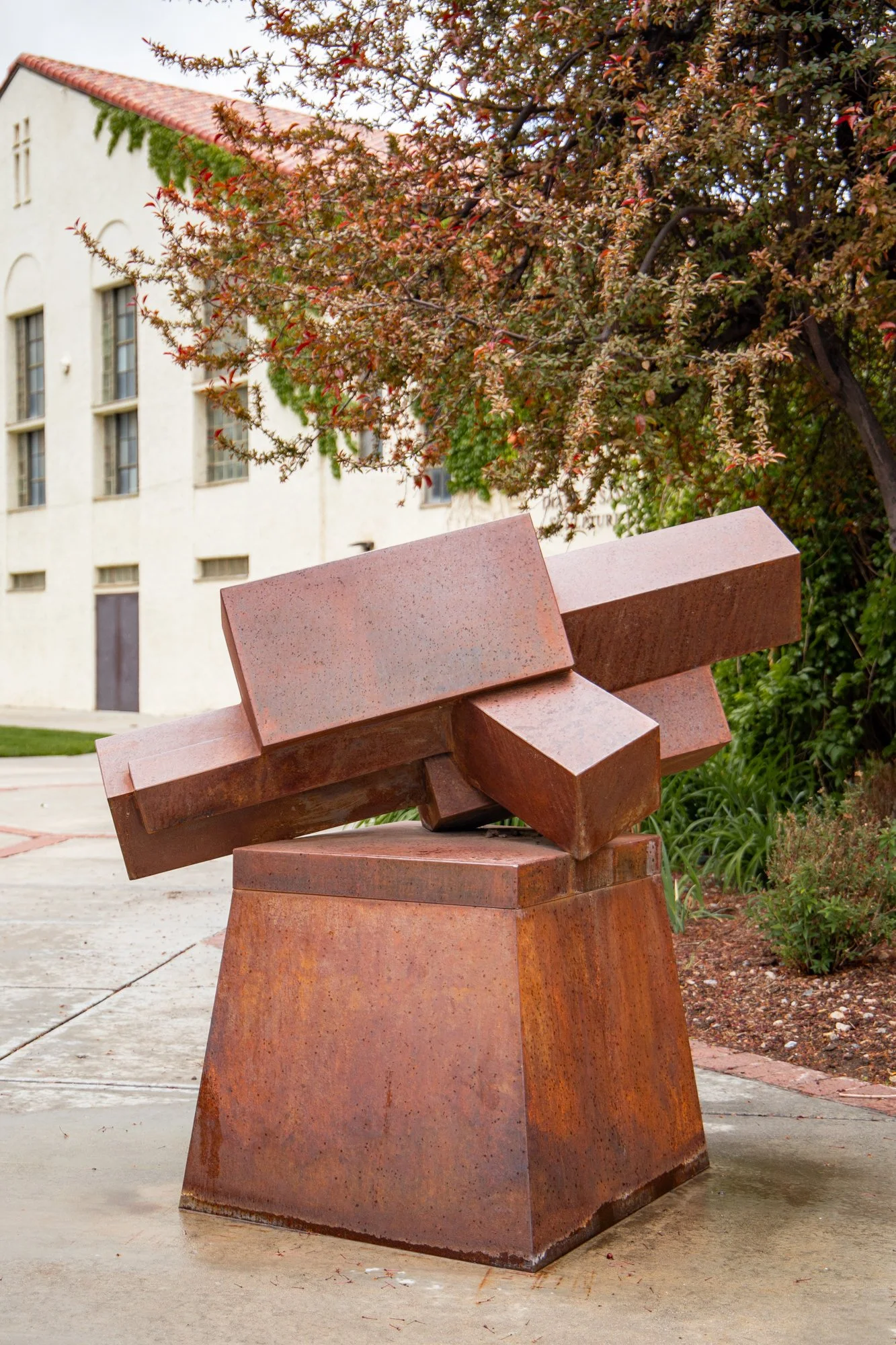A modern abstract sculpture made of rust-colored metal with geometric shapes, situated outdoors with trees and a building in the background.